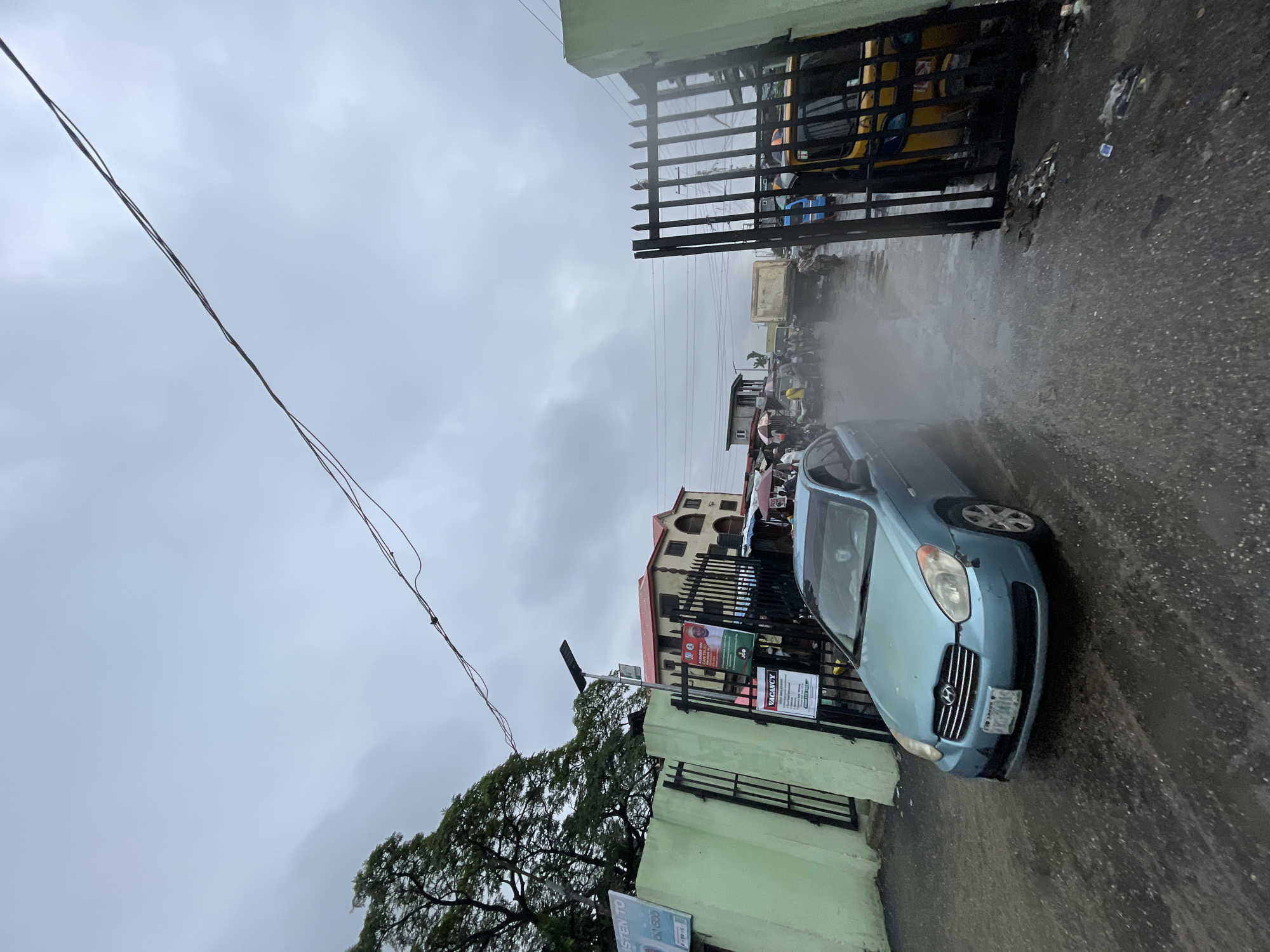 The image shows a street scene in Ado, Nigeria on a cloudy day, likely after or during rainfall given the wet asphalt. A light blue Hyundai Accent is parked near a gated entrance to a building, with the Nigerian license plate clearly visible. The building is painted in shades of green and cream. A power line runs across the upper left of the frame. In the background, behind a black gate, there's a yellow minibus and other indistinct vehicles and people suggesting a somewhat busy street scene. Debris and trash are scattered on the wet pavement near the gate. Political posters are affixed to the gate. The overall impression is one of a typical urban scene in Ado under overcast conditions.