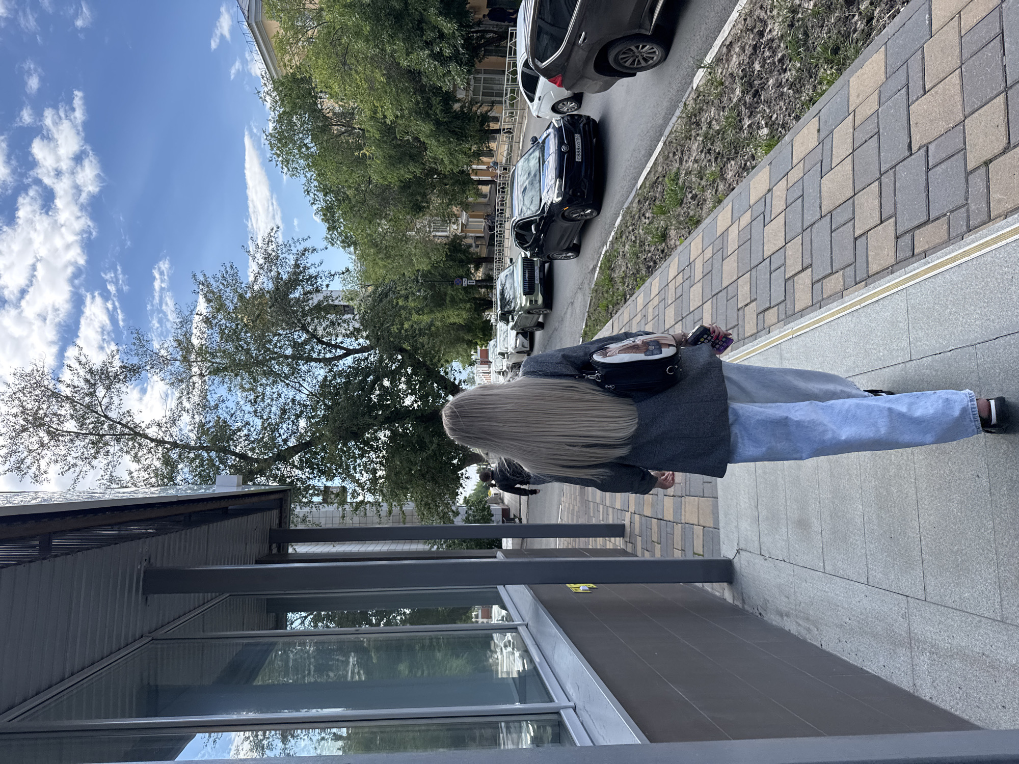 A person with long, blonde hair walks along a sidewalk in what appears to be a city setting. The person is wearing a grey jacket and light blue jeans. A black backpack with a picture on it is slung over their shoulder. The sidewalk is paved with rectangular tiles in shades of grey and beige. A street runs alongside the sidewalk, with several cars parked and driving on it. Trees with green foliage line the street and sidewalk. In the background, buildings are visible. The sky is blue with scattered white clouds, suggesting it is daytime and fair weather. The scene is oriented vertically in the image.