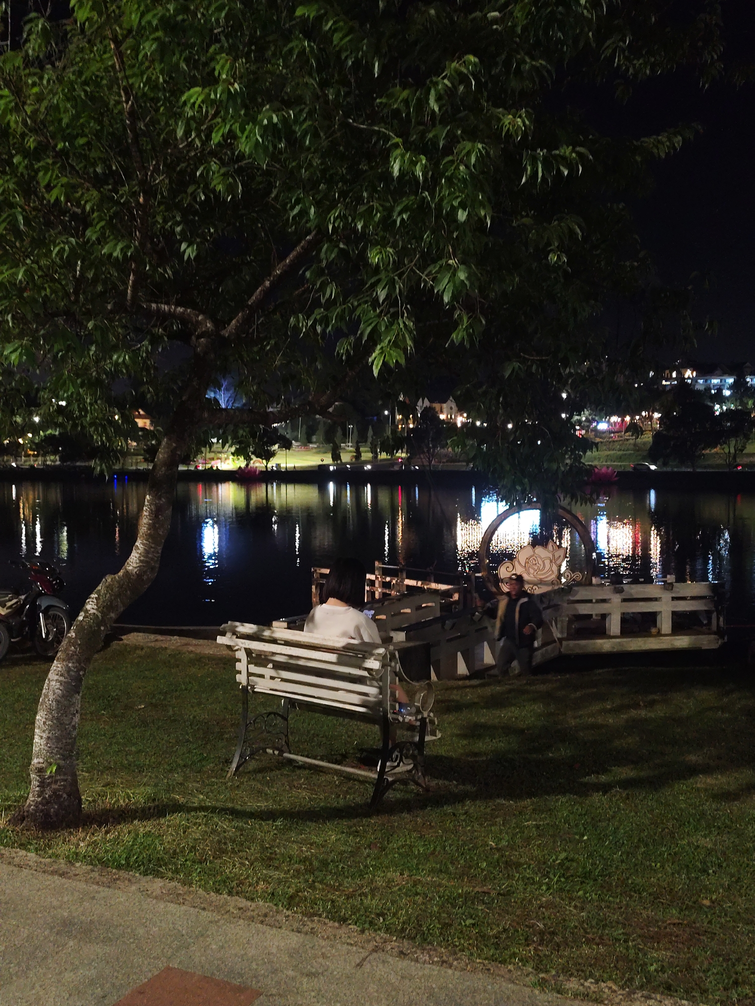 The image captures a nighttime scene in Da Lat, Vietnam, near Xuan Huong Lake. A person is seated on a white bench under a large tree, facing away from the viewer, looking toward the lake. The lake reflects the lights from buildings and decorations on the opposite shore. A motorcycle is parked on the left, and a decorative structure is positioned on a platform over the water on the right, with a person standing nearby. The surrounding area consists of a grassy lawn adjacent to a paved walkway.