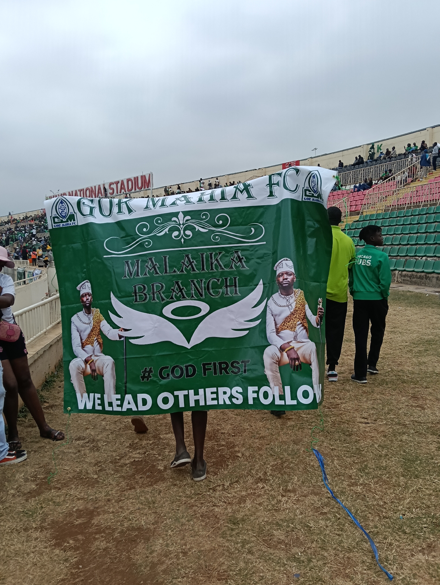 The image depicts a group of people at a stadium, likely during a sporting event. In the foreground, a large green banner is being held up, featuring images of two men in ceremonial attire. The banner displays text that reads "GOR MAHIA FC", "MALAIKA BRANCH", "# GOD FIRST", and "WE LEAD OTHERS FOLLOW". Behind the banner, spectators are visible in the stadium seating, which appears to be predominantly green and red. The stadium architecture suggests an outdoor venue with a partially cloudy sky. Based on the "GOR MAHIA FC" branding, the location is likely the Moi International Sports Centre (MISC) Kasarani in Nairobi, Kenya, which is the home stadium for the Gor Mahia Football Club. The ground surrounding the pitch is covered in dry grass. Several individuals are seen standing or walking in the vicinity of the banner, dressed in casual clothing and team apparel, including one person wearing a green jacket with "CHICAGO CUBS" printed on the back.