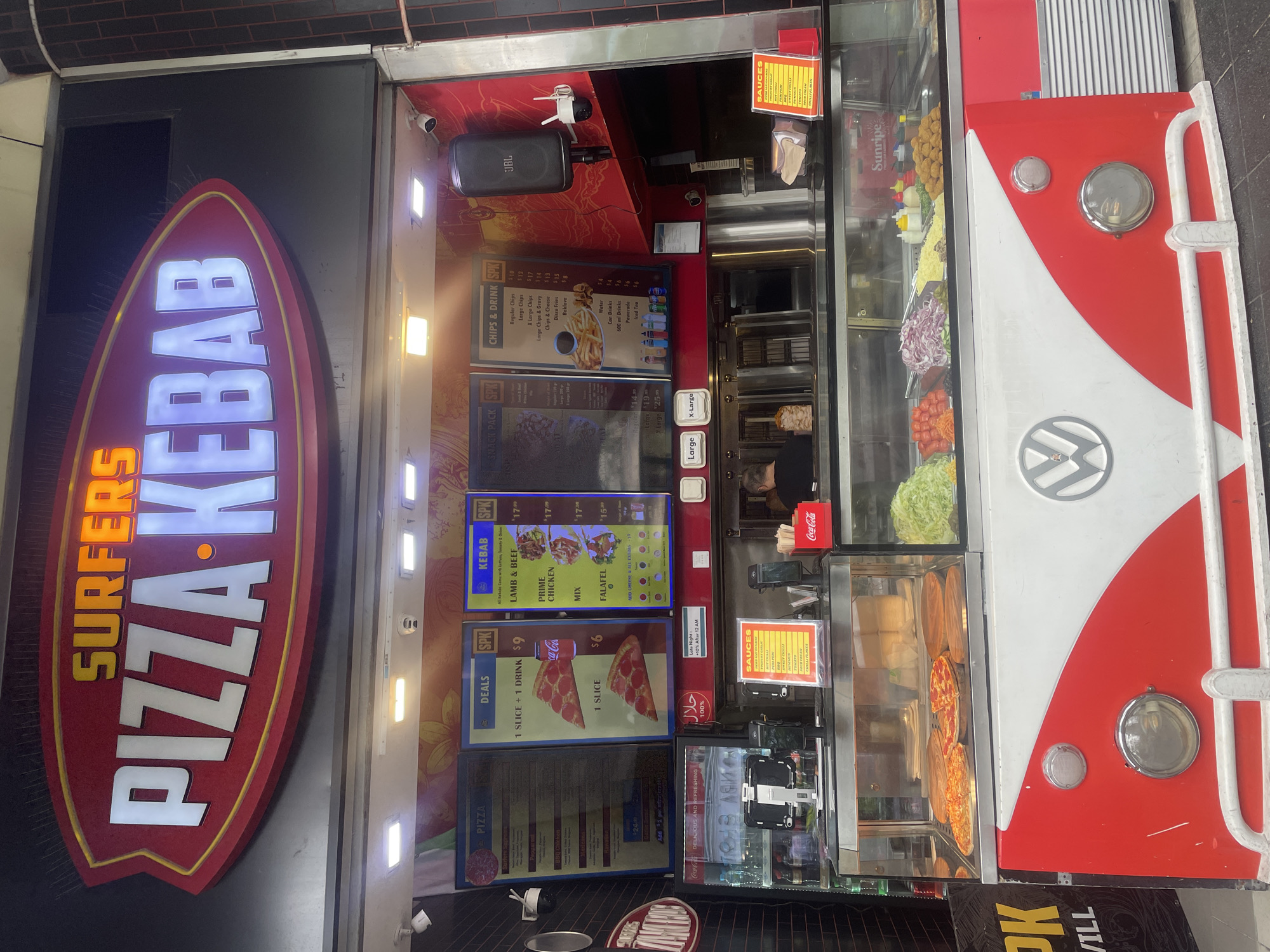 This image shows the exterior of "Surfers Pizza Kebab," a fast-food establishment located in Surfers Paradise, Australia. The facade of the shop features a prominent, brightly lit sign that reads "SURFERS PIZZA KEBAB." Below the sign, a display window showcases various food items, including pizzas, salads, and what appear to be kebab ingredients. The shop is designed with a retro, red and white color scheme, reminiscent of a Volkswagen van. There are menus displayed on the wall above the food display, detailing prices for pizza, kebab, chips, and drinks. In the foreground, the lower part of the building is decorated to resemble the front of a classic van, complete with headlights and a grille. A Coca-Cola cooler is visible to the right. Inside, a person can be seen working behind the counter, preparing food. The overall scene depicts a busy takeaway food outlet in a commercial area.