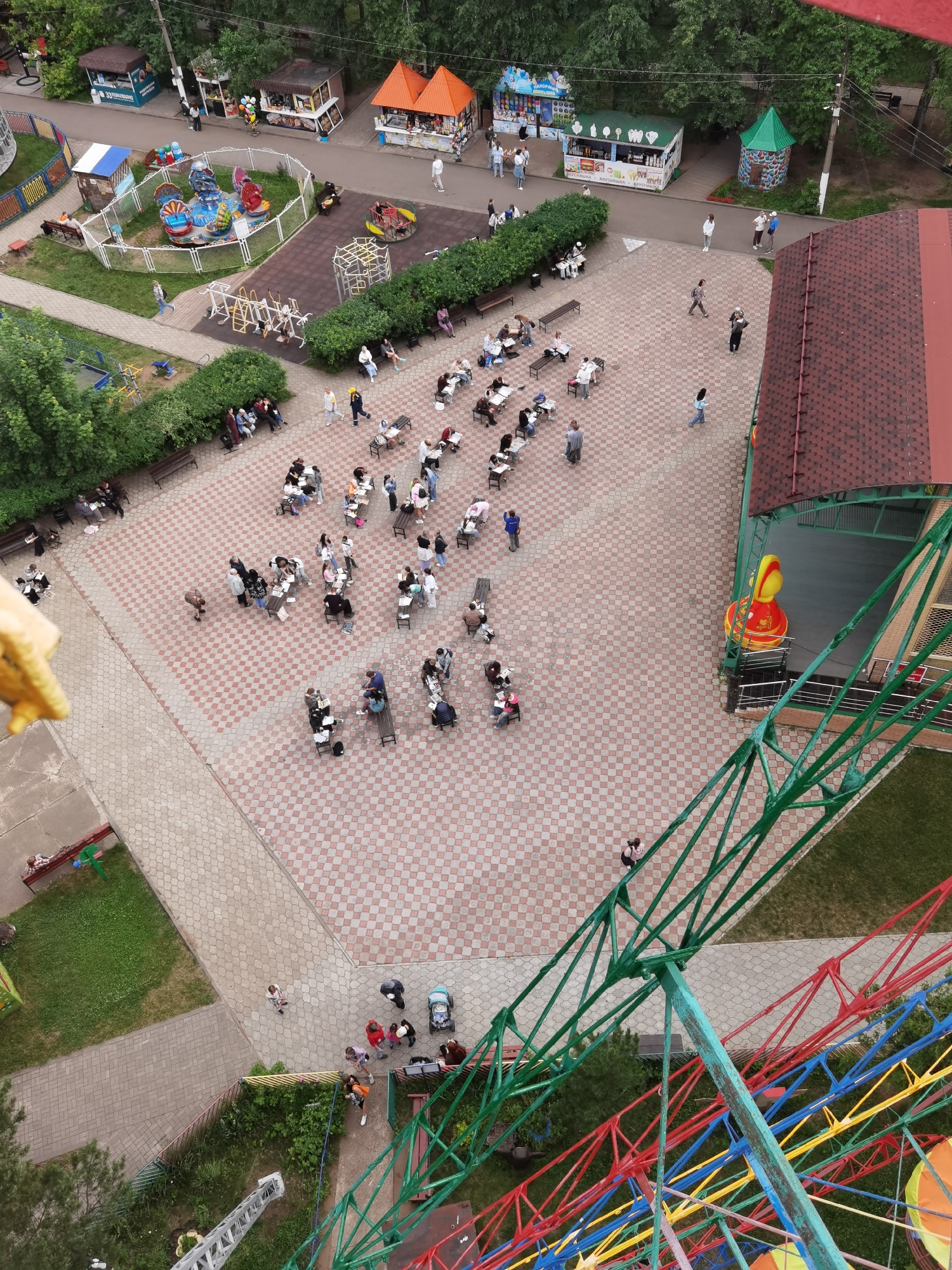 An aerial, high-angle shot captures an outdoor gathering in a park-like setting, likely in Chaikovsky, Russia. The scene is dominated by a large, checkered paved area where numerous people are seated at tables or on benches, engaged in activities that suggest a class or organized event, possibly related to art or education due to the presence of papers and pens. Surrounding this central area are green spaces with trees and bushes, interspersed with small market stalls or kiosks. In the foreground, a portion of a Ferris wheel structure is visible, with its colorful metal beams and framework extending upwards. A building with a red, tiled roof is also present on the right side of the image. The weather appears overcast.