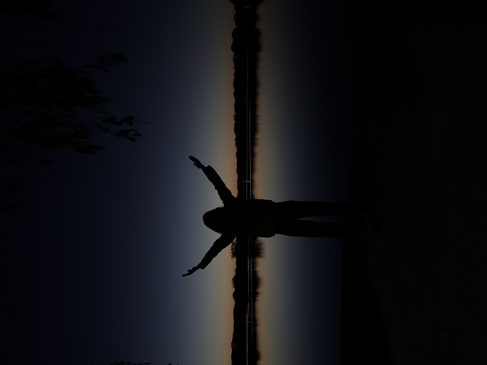 A silhouette of a person with outstretched arms is captured against a twilight backdrop. The background reveals a body of water reflecting a gradient sky, transitioning from dark blue to orange. A line of trees or foliage can be seen mirroring on the water’s surface. The darkness of the silhouette contrasts sharply with the dim light of the sky. The scene appears tranquil, possibly taken near a body of water in Haidian District, China, although specific details about the location are difficult to ascertain from the image alone.