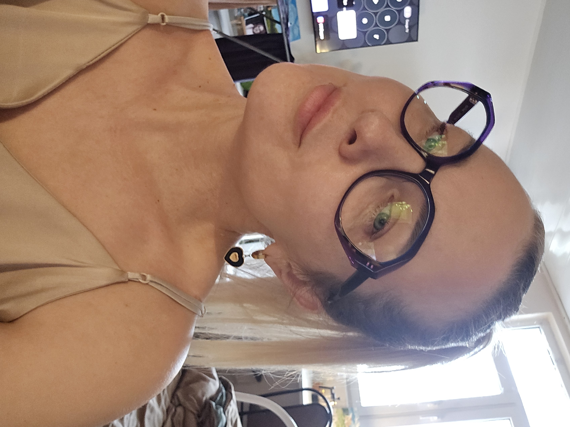 A fair-skinned woman with blonde hair, wearing eyeglasses, is the primary subject of this indoor photograph. She is wearing a light-colored top with thin straps. An earring is visible on her left ear. In the background, a television screen displays circular icons. Further back, a window and some furniture are visible. The location in the city of Sochi, Russia is not discernible from the visual information provided in the image.