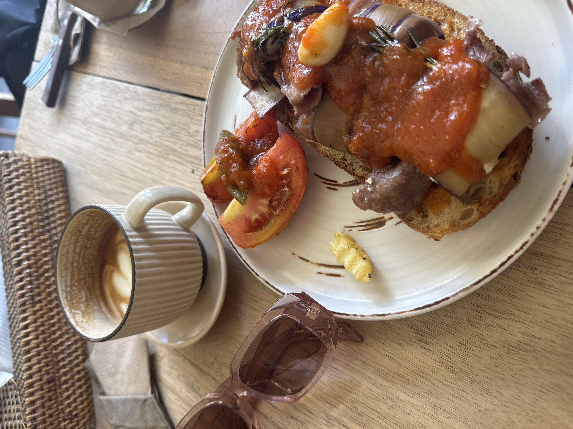 The image shows a partially consumed latte in a ribbed ceramic cup, a plate with an open-faced sandwich topped with what appears to be sliced eggplant, meat, and a tomato-based sauce, alongside a sliced tomato, and a single fried potato wedge. A pair of pink sunglasses lies beside the plate on a light-colored wooden table. A wicker basket is partially visible in the bottom left corner. The location is a restaurant in the city of Unknown, Unknown.