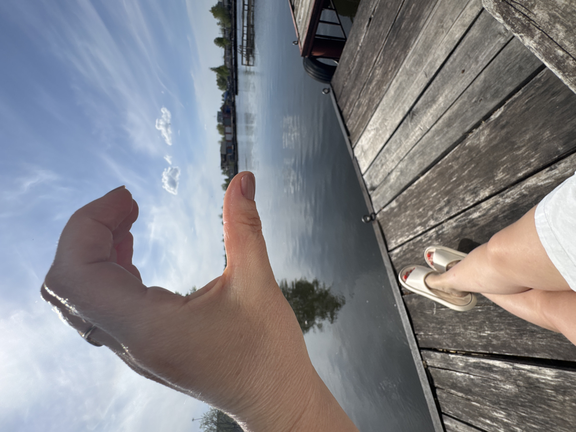 The image shows a hand held up against a backdrop of a waterway and sky. The person is likely sitting on a wooden dock or platform. The foreground features the hand, with the sky visible through the gap between the fingers. The waterway, which appears to be a river or canal, reflects the surrounding trees and sky. A portion of a building or structure is visible on the dock's edge. In the background, there are other buildings or structures along the waterway. There are no other indications of the city of Chaikovsky, Russia. The position of the camera suggests it was held by the person sitting on the dock. The image captures a moment of leisure and connection with nature.