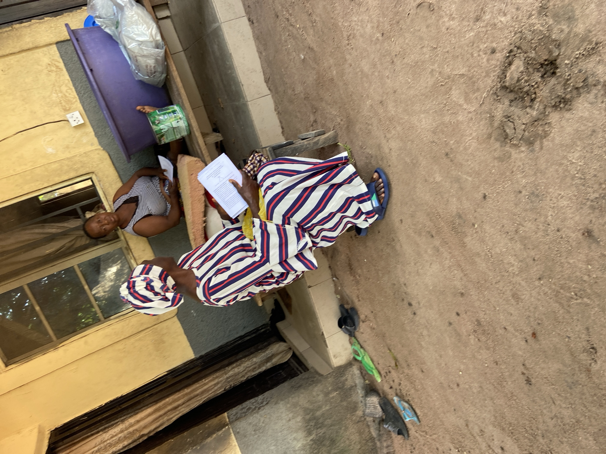 In the city of Mararaba, Nigeria, two women are seated outdoors on a low, wooden bench against a textured, yellow building. A woman in the foreground, wearing a white, blue, and red striped outfit and a matching headscarf, is holding a white document. She is seated on the left side of the bench and is looking down at the document. To her right, a second woman, wearing a black and white striped top, is also seated on the bench and holding a white document. She is looking towards the camera with a neutral expression. Behind the women, a large, purple plastic tub is visible, along with a white plastic bag containing what appears to be laundry. The ground in front of the bench is unpaved and consists of dirt and scattered debris, including a few pairs of footwear. A window with dark curtains is present on the building behind the seated women. The scene suggests a moment of rest or discussion outdoors.