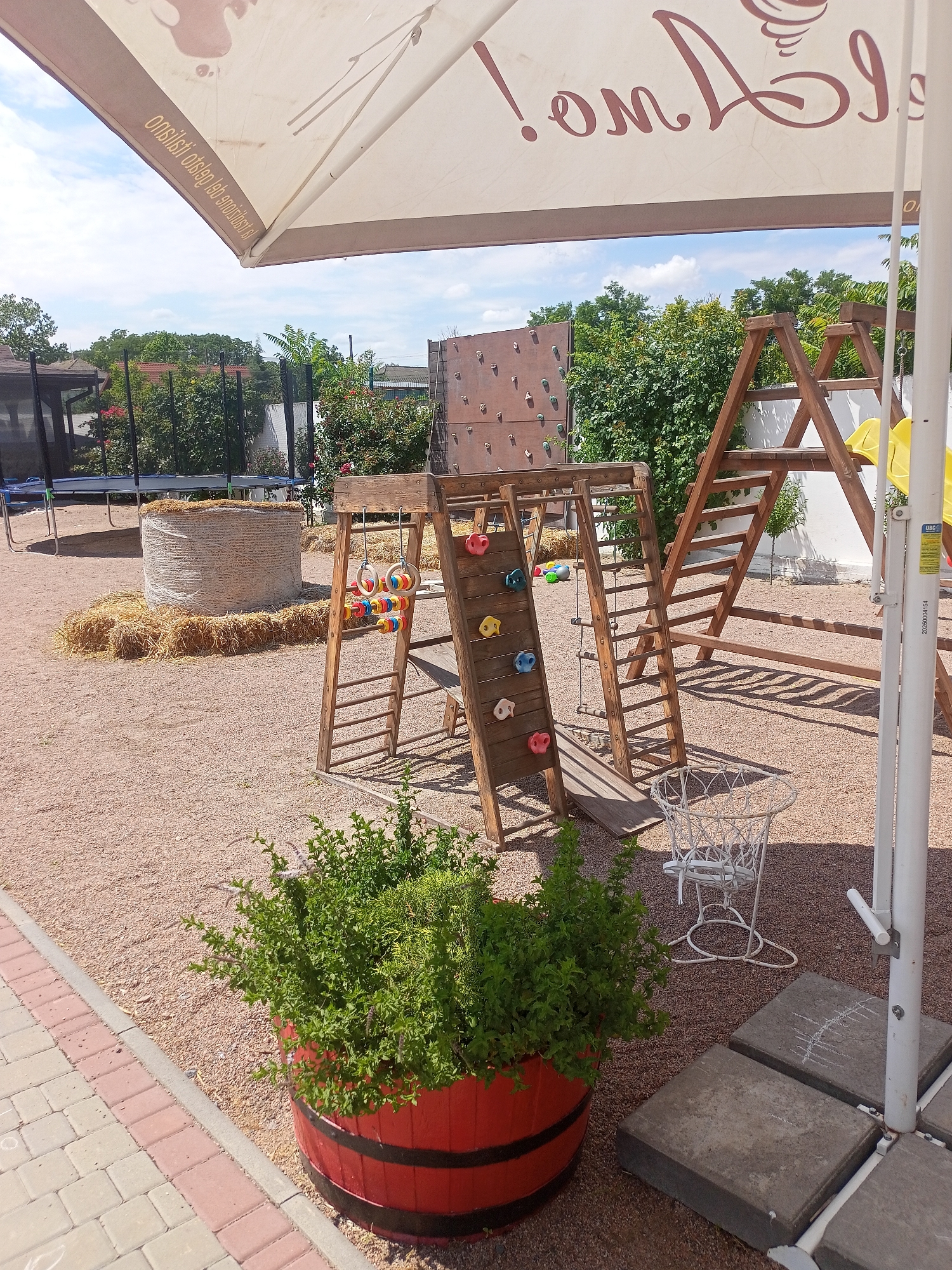 An outdoor recreational area in the city of Shabo, Ukraine, features a wooden playground structure with a climbing wall, ladder, and slide. Nearby, a large bale of hay sits on a bed of straw, with a trampoline visible in the background. A red barrel planter filled with green bushes adds a decorative element. The ground is covered in gravel, and a paved walkway lines the edge of the area. A large white umbrella provides shade, and a white metal basket-like object stands near the playground. The scene is set against a backdrop of lush green trees and a partly cloudy sky.