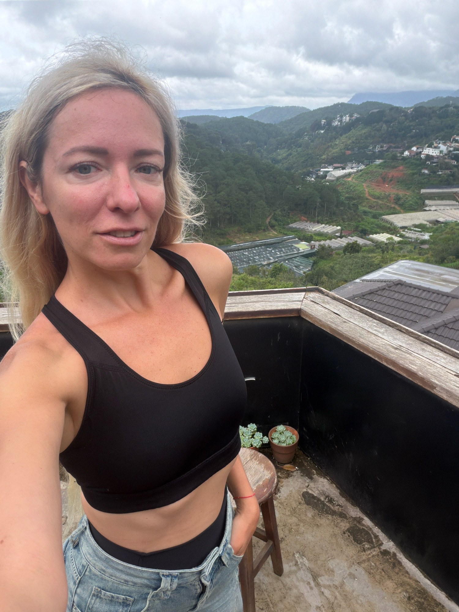 A Caucasian woman with blonde hair is taking a selfie on a balcony overlooking a valley in Da Lat, Vietnam. She is wearing a black sports bra and denim shorts. The background shows rolling hills covered in green vegetation, with some buildings and greenhouses visible in the distance. The sky is overcast.