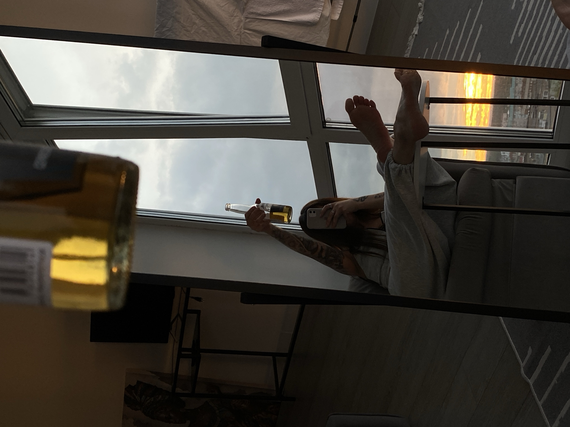 The image is a selfie taken by a person lying down, with their legs propped up and visible in the foreground. They are holding a bottle of liquid, possibly beer or wine, and are looking at a phone, presumably taking the photo. The person has tattoos on their arm and is wearing casual clothing.
The scene is likely indoors, near a large window or glass door that offers a view of the sky. The sky appears overcast with some hints of sunset or sunrise light in the distance. The reflection in a mirror or glass surface dominates the image, showing the person and the window.
The overall mood is relaxed and perhaps contemplative, suggested by the leisurely pose and the drink. The lighting indicates it is either early morning or late evening. There is no visible text other than what might be on the label of the bottle, which is partially obscured and unreadable.