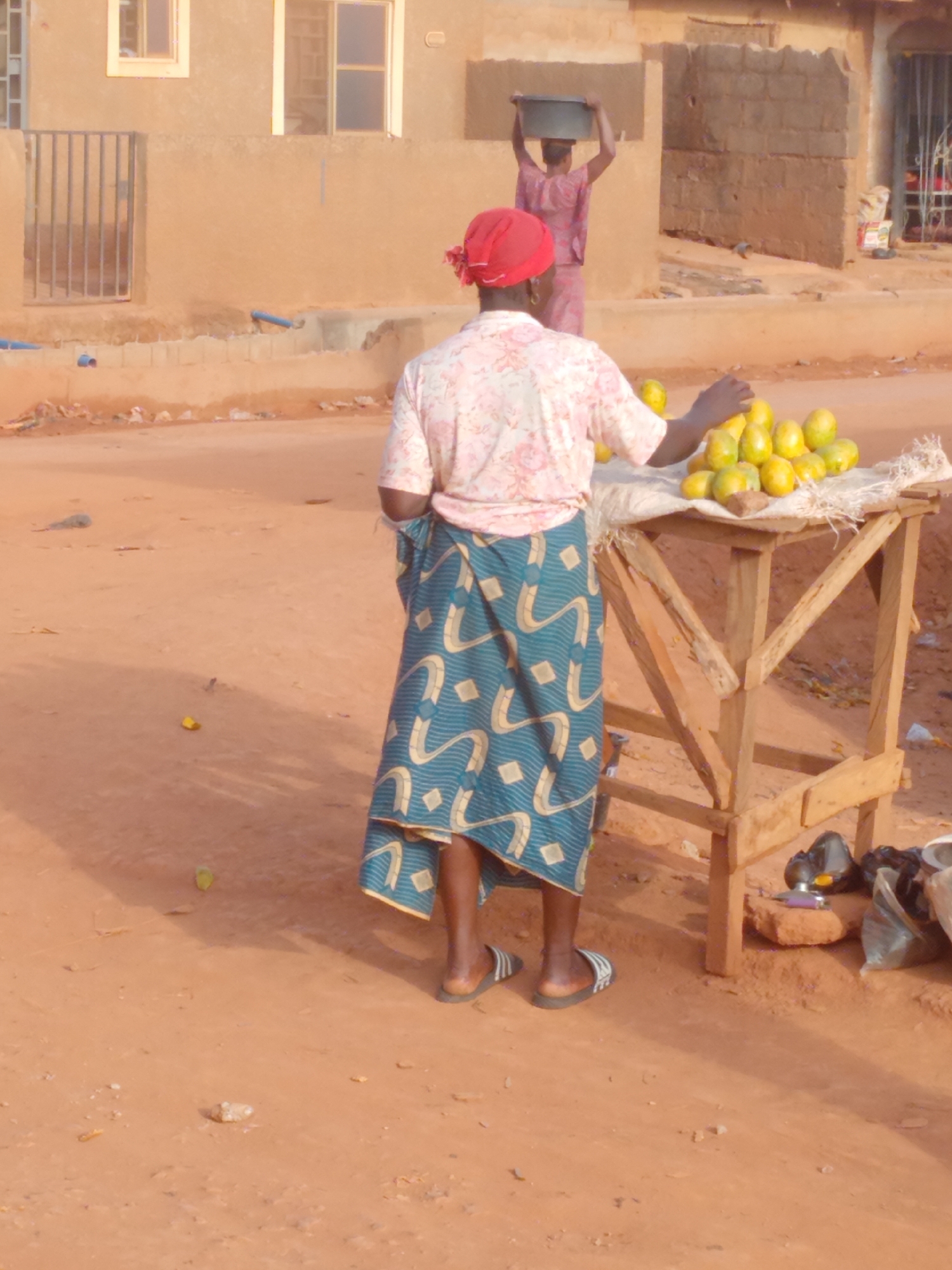 This outdoor photograph captures a bustling scene of daily life in Yelwa, Nigeria, featuring two individuals engaged in everyday activities on an unpaved, reddish-brown dirt road. The overall impression is one of local commerce and daily routines under bright daylight.
In the foreground, viewed from behind, stands a woman who appears to be a street vendor. She is dressed in a light-colored, short-sleeved top with a subtle floral pattern, paired with a vibrant blue wrap skirt adorned with light-yellow geometric designs. A red head covering completes her attire, and she wears black and white striped flip-flops. Her right hand rests near a simple wooden stall, draped with a white cloth, on which a bountiful pile of green and yellow fruits, likely mangoes or papayas, is displayed for sale. Beneath the stall, various items are visible, including black plastic bags and a small, colorful object resting on a rock. Several fruits are scattered on the ground near the vendor's feet.
Further in the background, another person, also seen from behind, walks away, carrying a dark basin or bowl balanced on their head – a common method of transport in the region. This individual is dressed in a light pink or purple patterned outfit.
The surrounding environment consists of buildings constructed from unpainted or lightly rendered mud/concrete blocks, some featuring windows with metal grills. Blue pipes are visible on the ground to the left, near one of the buildings. The bright, clear lighting indicates a daytime setting, likely mid-morning or late afternoon, as suggested by the moderate shadows. No visible text is present in the image.