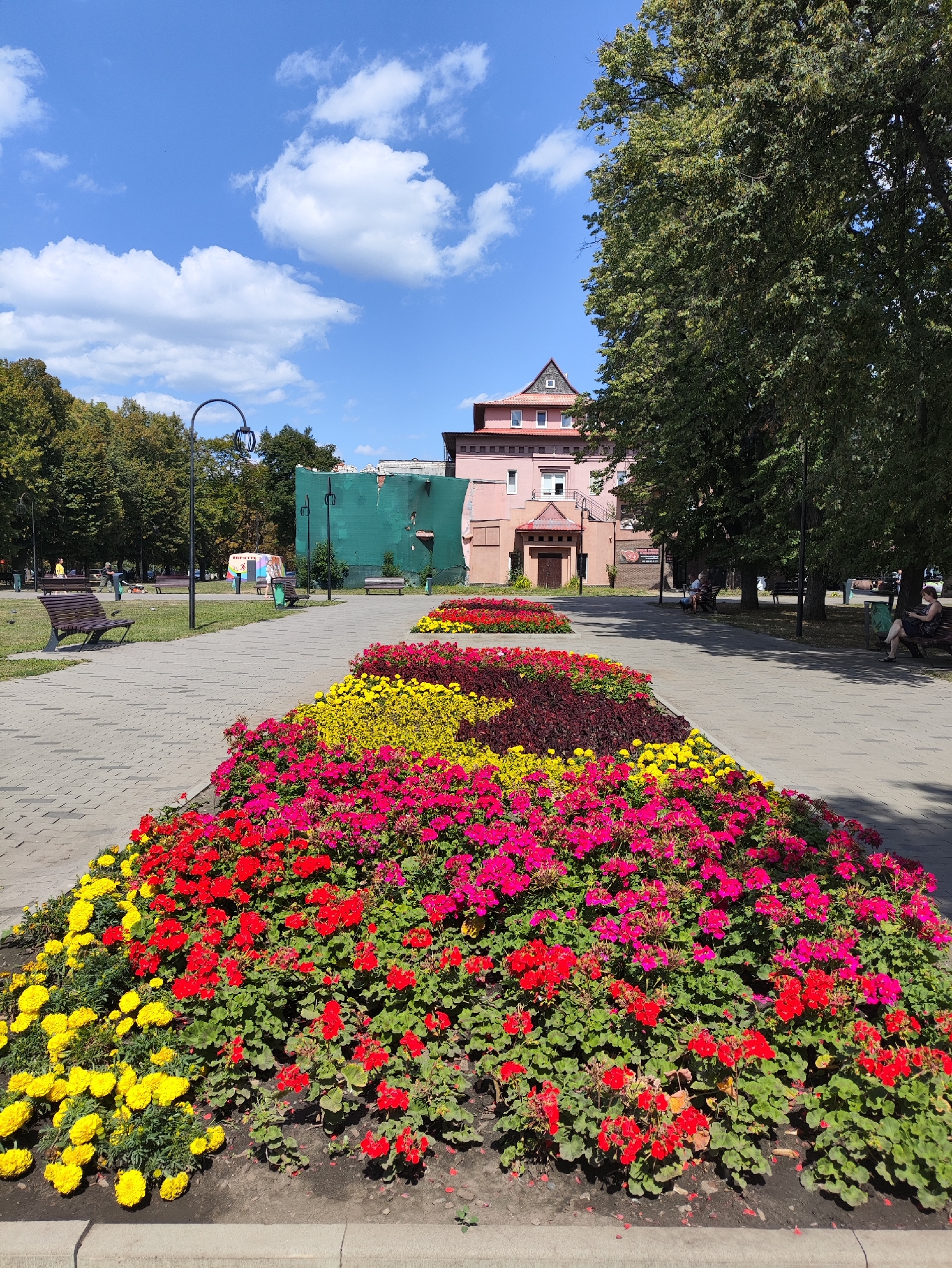 The image depicts a sunny day in a park in Kharkiv, Ukraine. In the foreground, a long, raised flower bed is filled with vibrant red, pink, and yellow flowers. A paved walkway runs alongside the flower bed, with wooden benches placed at intervals. In the background, trees line the park, and a pink building with a pitched roof is visible. A large, green, tarp-covered structure is also present, possibly under construction or renovation. People are seen sitting on benches and walking along the path. The sky is blue with scattered white clouds.