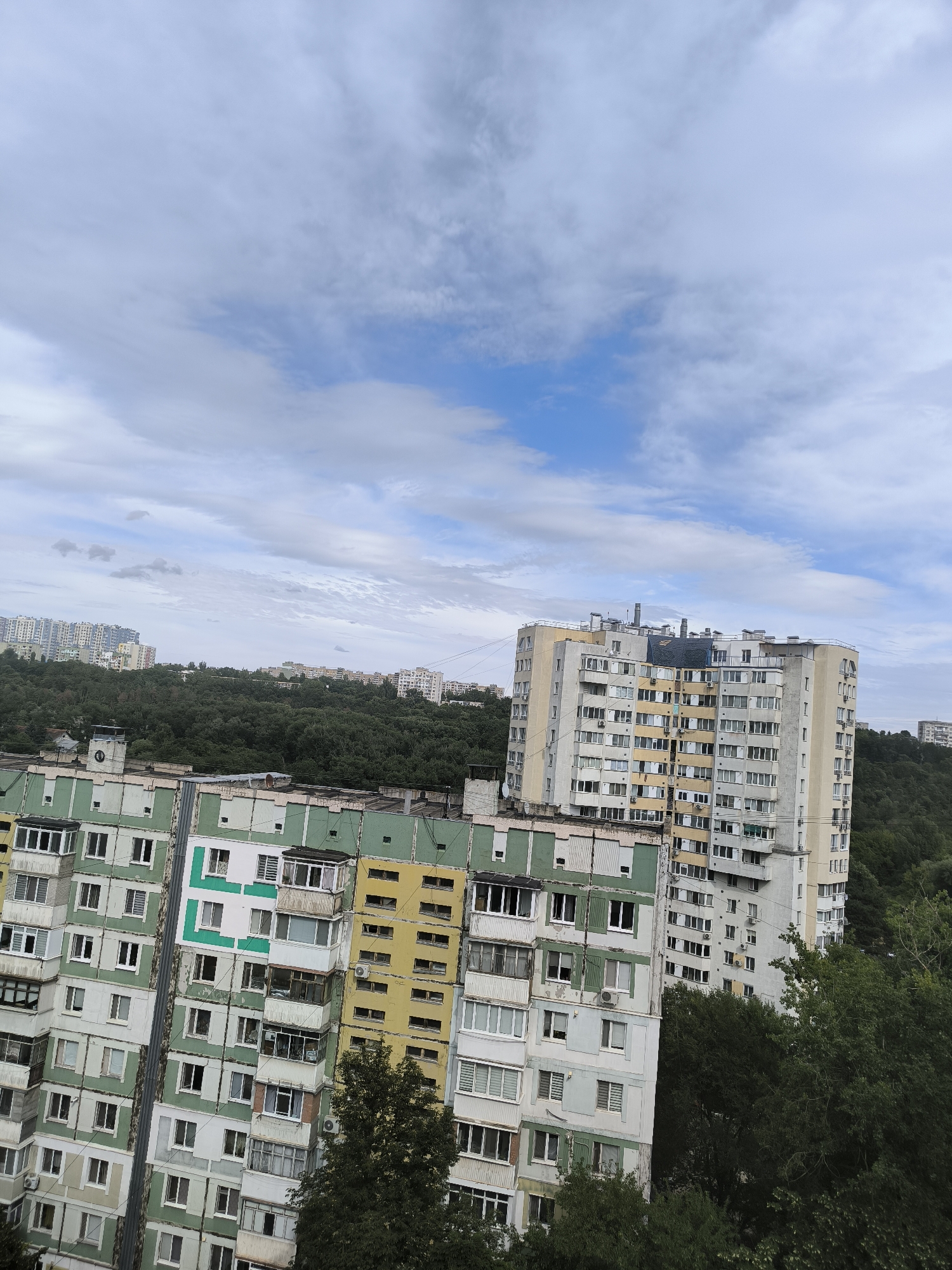 The image shows a panoramic view of a residential area in Chișinău, Moldova. The foreground is dominated by the facades of several large, multi-story apartment buildings. These buildings exhibit a variety of architectural styles and color schemes, with some featuring a patchwork of green, yellow, and white sections, while others are primarily gray and cream colored. Numerous windows and balconies are visible, some with air conditioning units attached.
In the mid-ground, a dense, green forest covers a hillside, separating the foreground apartment blocks from more distant buildings. These further buildings appear to be more modern, higher-rise apartment complexes, suggesting a mix of older and newer developments within the city. The background reveals a cloudy sky with patches of blue, indicating a daytime scene with somewhat overcast weather. The overall impression is of a densely populated urban environment interspersed with natural green spaces. No people are clearly visible, and no text is discernable in the image. The image conveys a sense of everyday urban life.