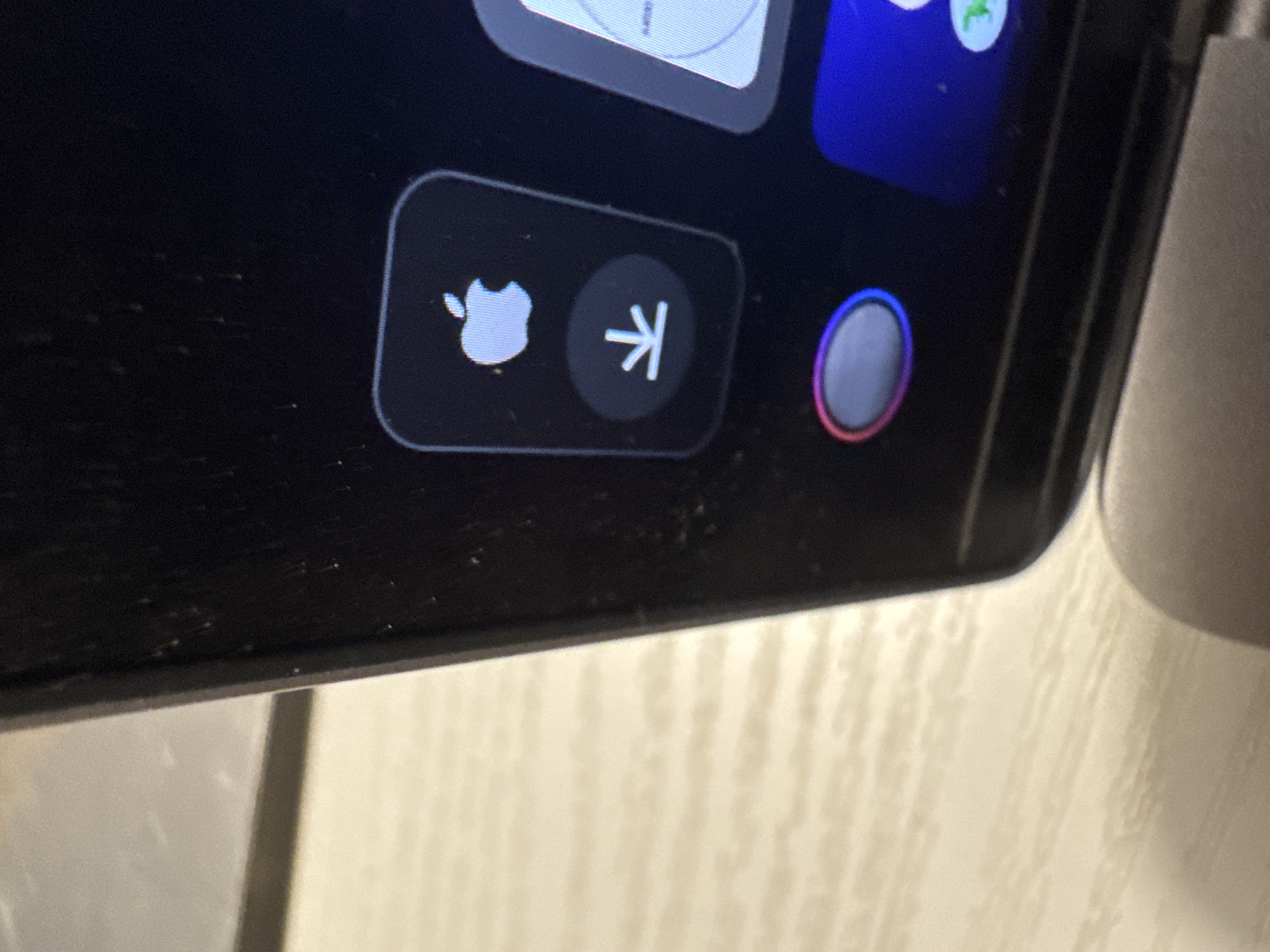 A close-up shot shows a dark, rectangular screen, likely a smartphone or tablet, displaying icons. On the left, within a rounded rectangular button, is a white Apple logo, slightly bitten. To the right of the Apple logo is a white downward-pointing arrow icon within a circular button, and further to the right, a circular area with a gradient of colors, ranging from light gray to pink and purple, hinting at a user interface element. The screen appears to have dust or smudges on it. The device is positioned against a textured, light-colored surface, possibly a table or wall, in Kuta Selatan, Indonesia. The lighting suggests an indoor setting. No people are visible.