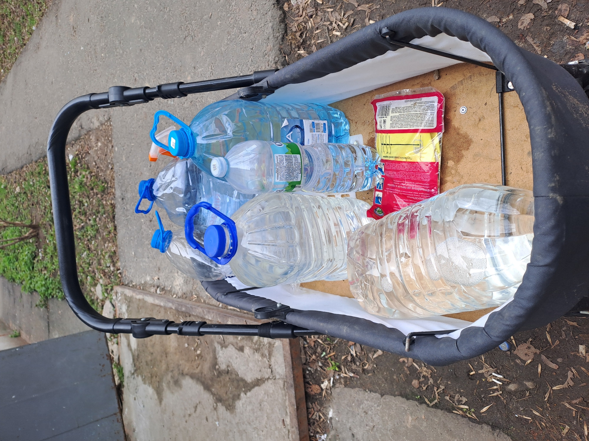 Here's a detailed description of the image:
**General Description:**
The image shows a top-down view of what appears to be a stroller or small cart filled with several large plastic water bottles and a package of gloves. The stroller is positioned on an outdoor surface that appears to be a sidewalk or area adjacent to a road.
**Objects:**
* **Water Bottles:** There are approximately 6-8 large plastic water bottles, mostly transparent, filled with clear liquid (presumably water). The bottles vary in size and shape, with some having blue caps and handles. Some of the bottles have labels.
* **Gloves:** A package of gloves with a red background is also in the stroller. Some of the text on the packaging is in Russian. The gloves are described as embossed on the fingers and palms.
* **Stroller/Cart:** The stroller or cart has a black frame and a lined container area. The interior appears to be a light color. The stroller is designed for carrying objects and is not designed for children.
**Environment:**
* **Outdoor Surface:** The surrounding environment consists of a concrete or asphalt surface. There is some vegetation (grass) and leaves visible in the background.
* **Location:** The presence of text in Russian on the glove packaging and analysis of a visual element (the gloves) suggests the location is likely in Elektrostal, Russia.
**Details:**
The scene appears to be utilitarian rather than staged. The collection of water bottles could imply a purpose such as recycling, storage or delivery. The presence of the gloves may indicate a task that requires hand protection.