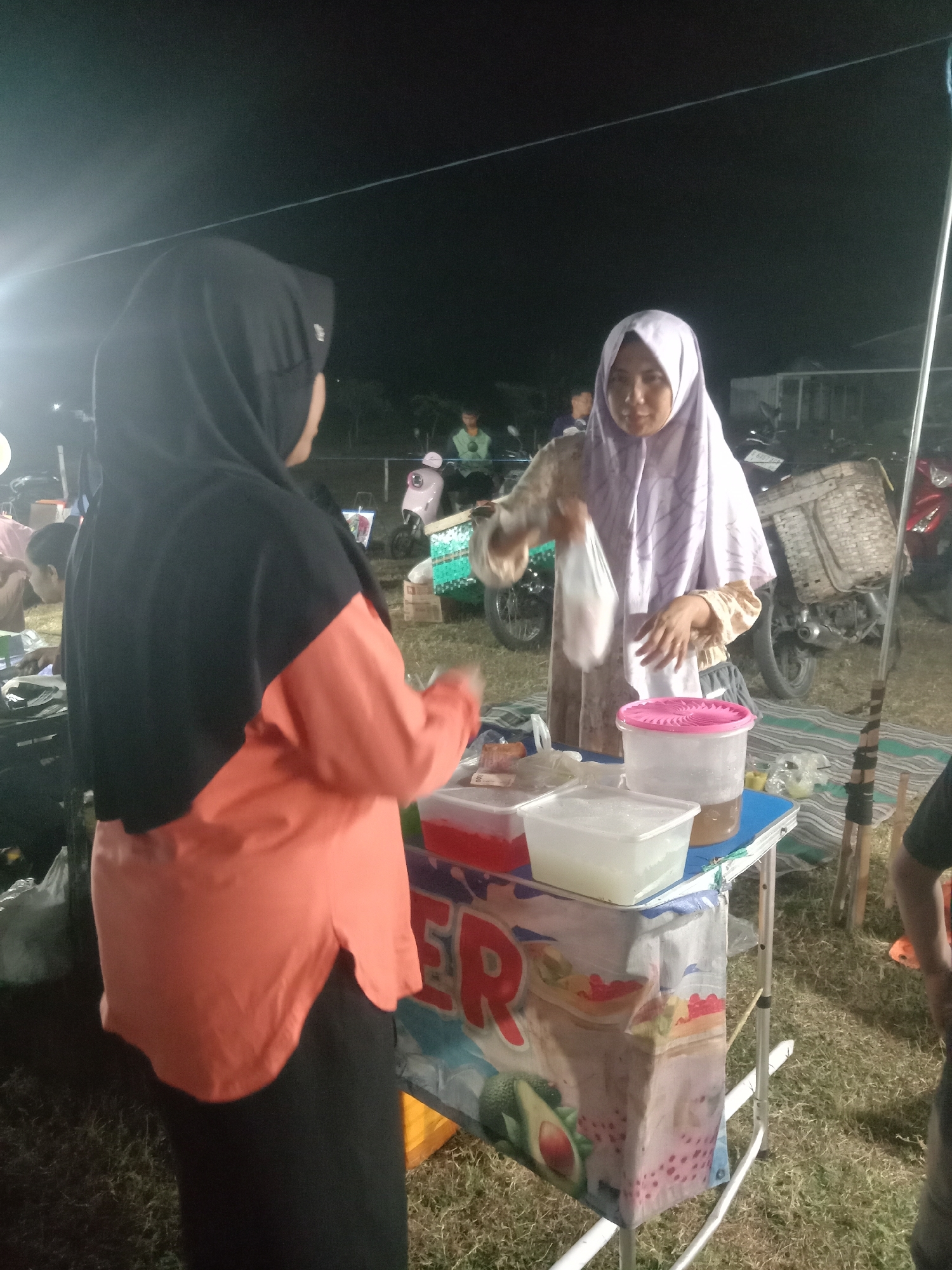 The image depicts an outdoor night scene at what appears to be a street market or fair in Stiaji, Indonesia. In the foreground, two women are interacting near a stall. The woman on the left, wearing a black hijab and an orange and black shirt, is partially visible from the back. The woman on the right, wearing a light purple hijab and a patterned shirt, is holding a clear plastic bag containing what seems to be food or a drink. In front of the woman on the right is a small table with various containers, including plastic tubs filled with liquids and possibly ice. A pink-lidded plastic container is prominent. The stall itself is decorated with a colorful banner displaying images of drinks and fruit. In the background, there are motorcycles and other people, suggesting a lively gathering. The ground is covered with grass and some striped mats. The overall atmosphere is that of a bustling marketplace at night.