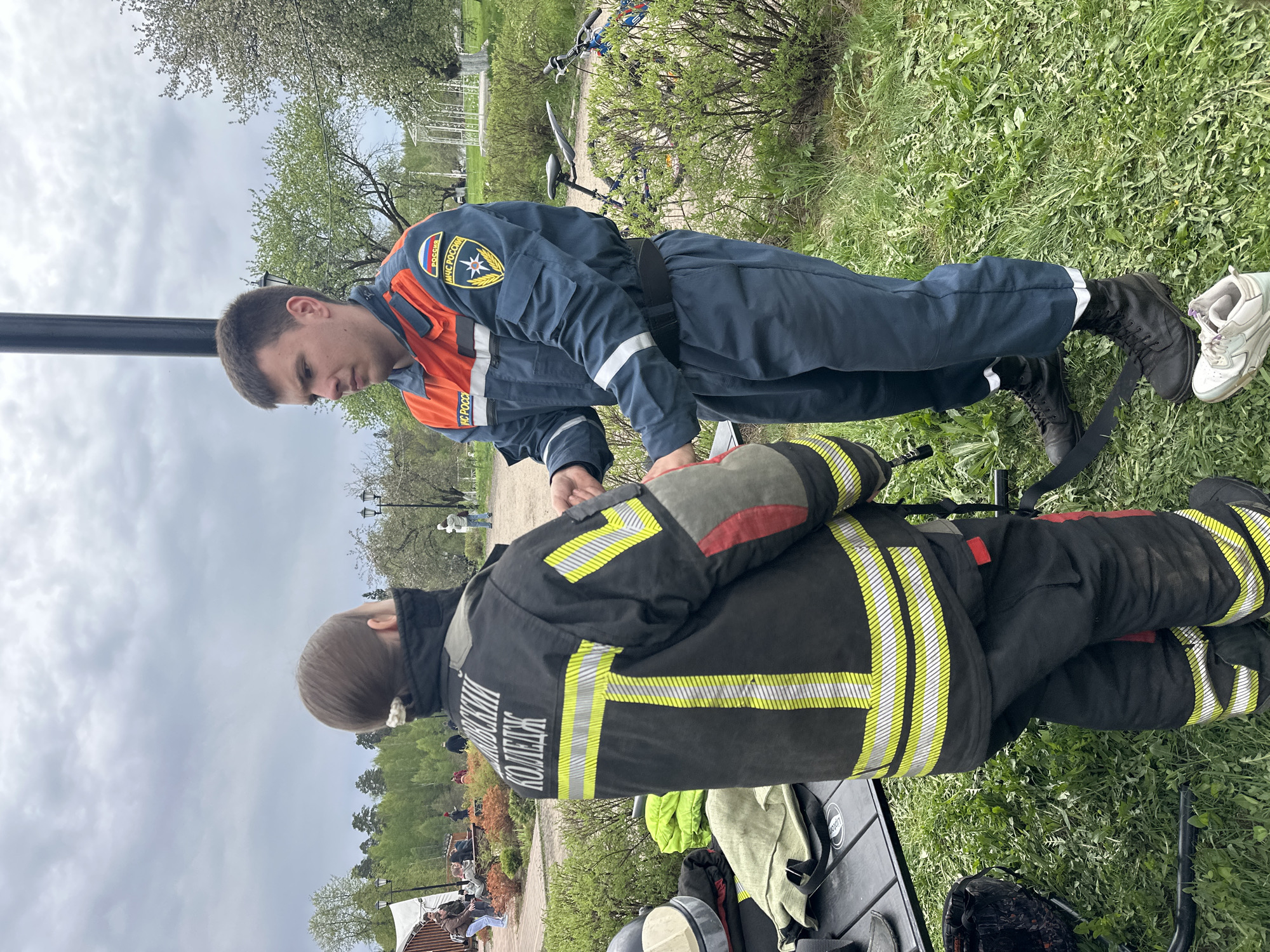 Two men are in a park setting in Grebnevo, Russia. One man is wearing a dark blue uniform with orange accents and patches indicating "МЧС России" and a symbol depicting a white starburst within a blue shield. He is bending over, looking intently at the other man. The second man is wearing a black firefighter's suit with yellow reflective stripes. The lettering on the back reads "МОСКОВСКИЙ КОЛЛЕДЖ". The men are outdoors on a grassy area, near a paved path, where people are sitting. Other objects are visible, including a table, a bicycle, and greenery.