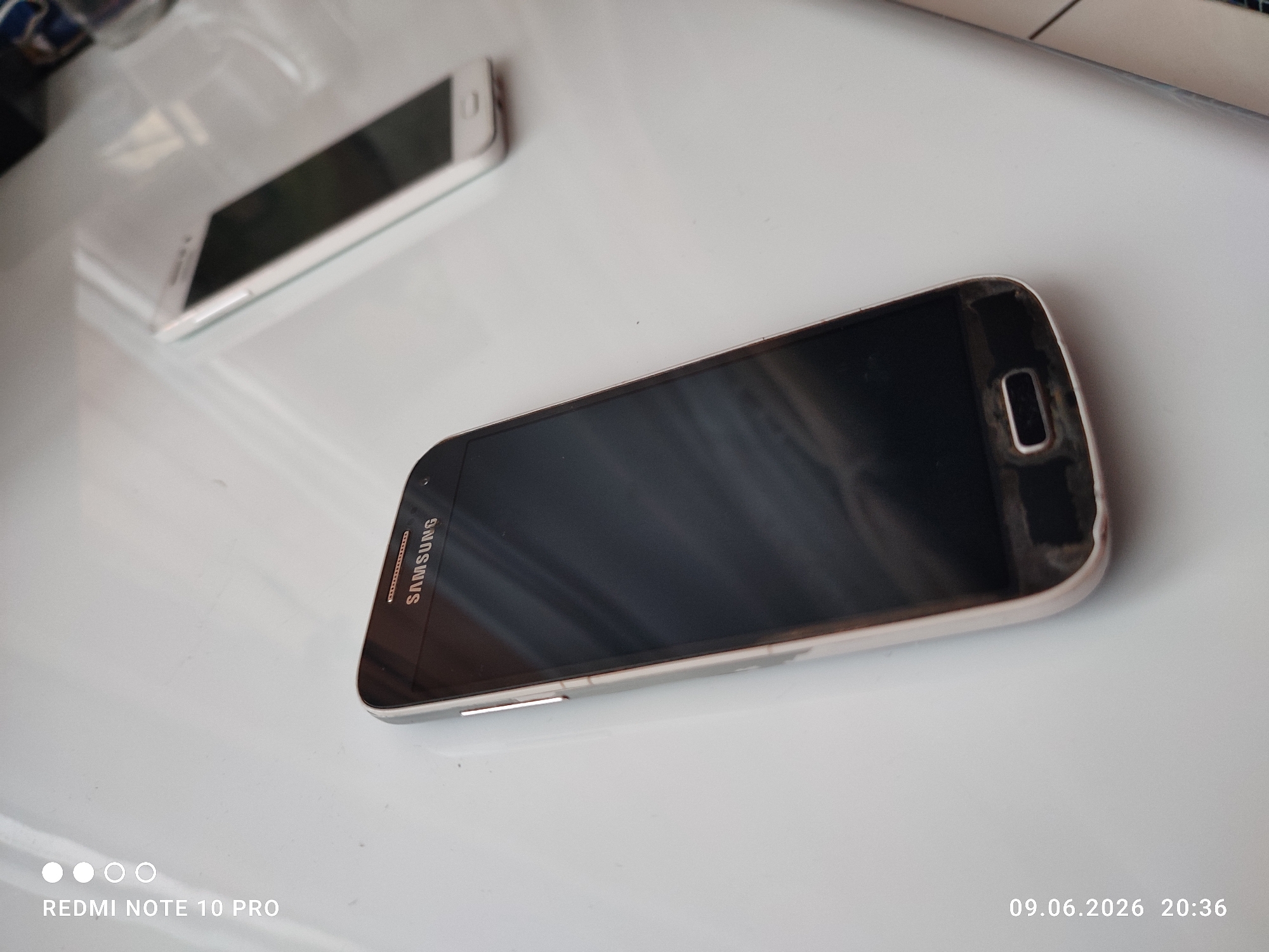 Two Samsung smartphones are visible on a white surface. One is positioned horizontally in the foreground, displaying its screen and the Samsung logo on its bezel. The other, smaller smartphone is positioned diagonally in the background, also showing its screen. The surface appears to be a desk or tabletop, with a faint reflection visible on its surface. The image is dated June 9, 2026, at 20:36.