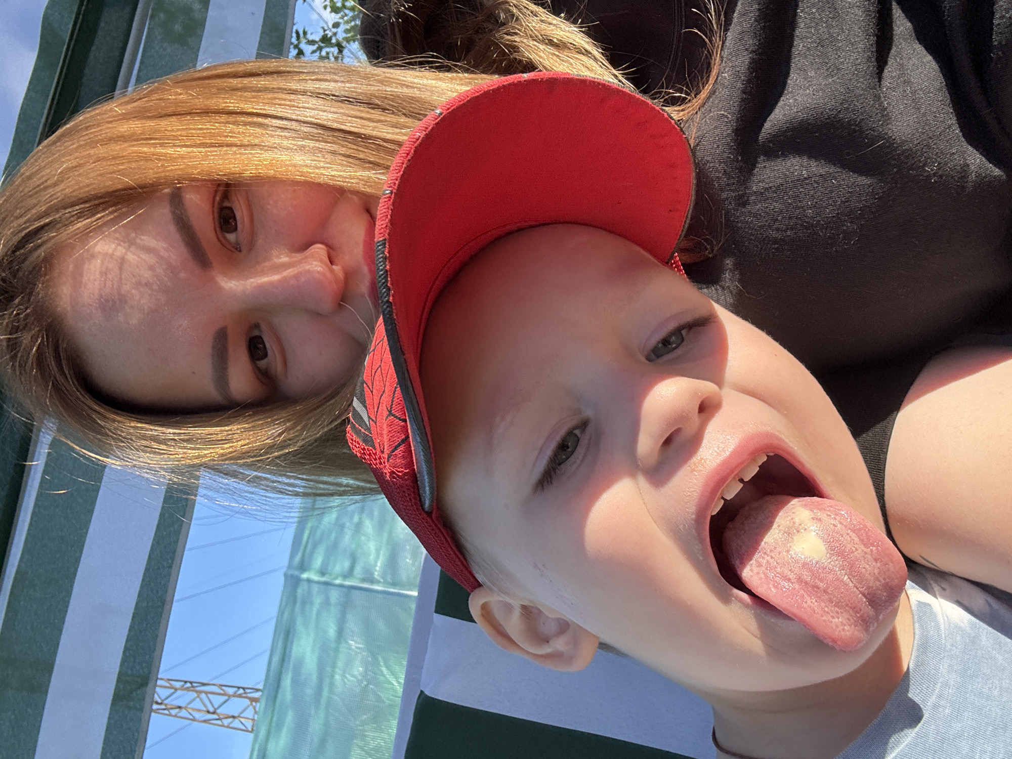 This is a selfie taken outdoors, likely on a sunny day, with two individuals. In the foreground, a young child with a red cap is sticking out their tongue. The child's eyes are visible, and they appear to be looking at the camera. Behind the child, a woman's face is partially visible. She has blonde hair and is smiling. The background shows a blue sky, some green netting, and a structure that resembles a power line tower. The overall setting suggests a casual, outdoor activity. The image appears to have been taken in Chaikovsky, Russia, given the context of the request, though there are no explicit visual cues within the image itself that confirm this location.