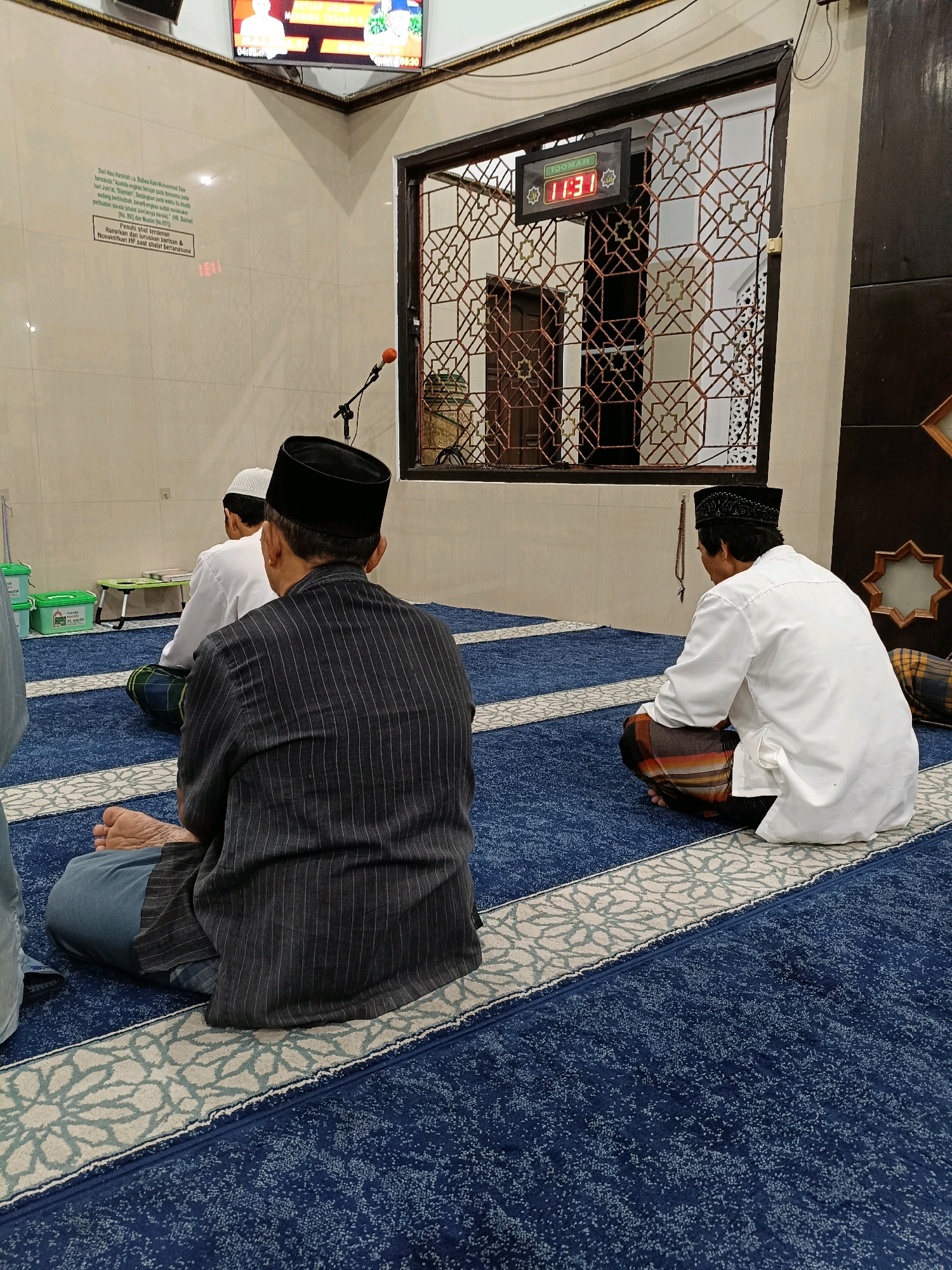 This image depicts the interior of a mosque in Maguwoharjo, Indonesia. Three men are seen sitting on a blue carpet with a patterned design. Two of the men are facing away from the camera, while one is facing towards the right side of the frame. All three are wearing traditional Islamic attire, including peci hats. One man in the foreground is wearing a dark pinstriped shirt and grey pants, while the man to his right is wearing a white long-sleeved shirt and a patterned sarong. Another man, further in the background, is wearing a white shirt and a white peci.
The mosque's interior features beige tiled walls and a decorative window with intricate metalwork. A digital clock displaying "11:31" is visible through the window. A microphone is positioned on a stand near the wall. In the background, there are green containers and a foldable table. A television screen is mounted high on the wall, displaying an image of people. The overall atmosphere suggests a moment of prayer or religious gathering.