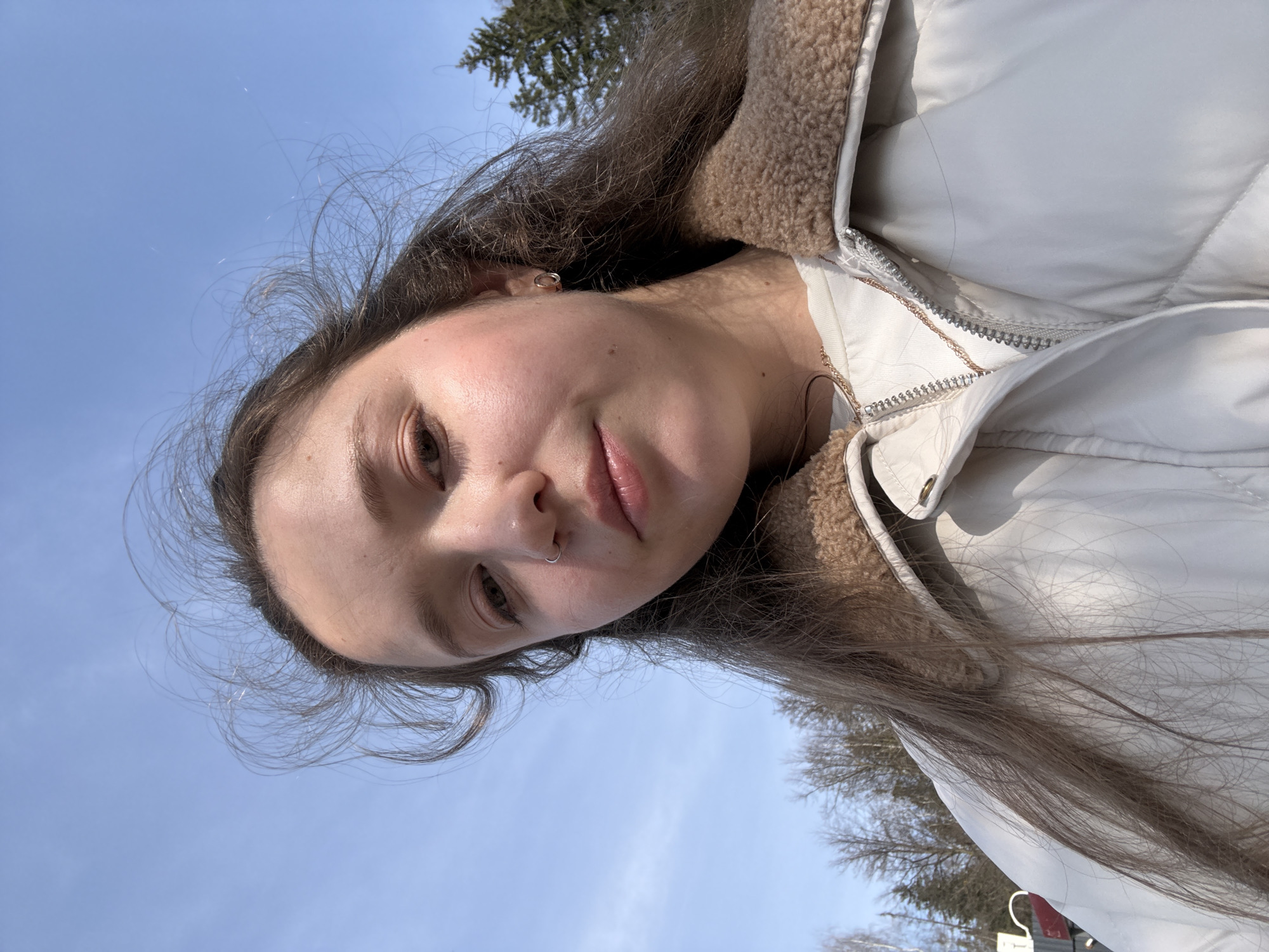 A selfie of a young woman taken from a low angle. She has long brown hair, fair skin, and is wearing a light-colored puffer jacket with a fur-lined collar. She has a nose ring. The background shows a clear blue sky and the tops of bare trees, indicating a possible winter or early spring setting. The location is indeterminate, there is no definitive indication that this is in Izhevsk, Russia.