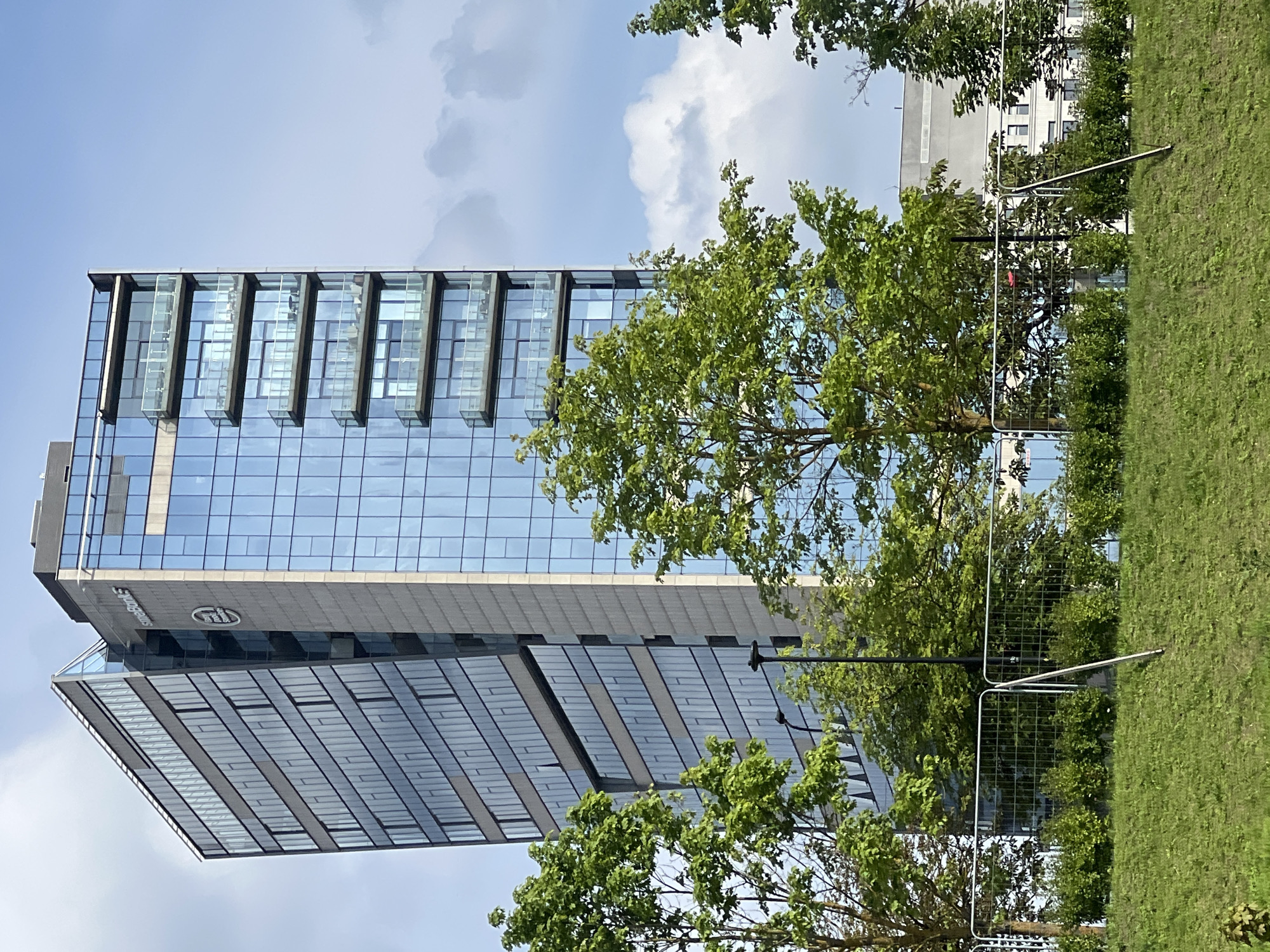 A modern, multi-story glass-fronted office building stands tall against a cloudy blue sky in Vilnius, Lithuania. The building features a unique architectural design with a slanted facade on the left side. Several floors of the building are visible, showcasing balconies with glass railings and reflective blue windows. The building is partially obscured by lush green trees in the foreground. A metal fence runs horizontally across the lower part of the image, with a grassy slope in front of it. Another, less distinct building can be seen in the background. The scene appears to be an exterior shot on a bright day.