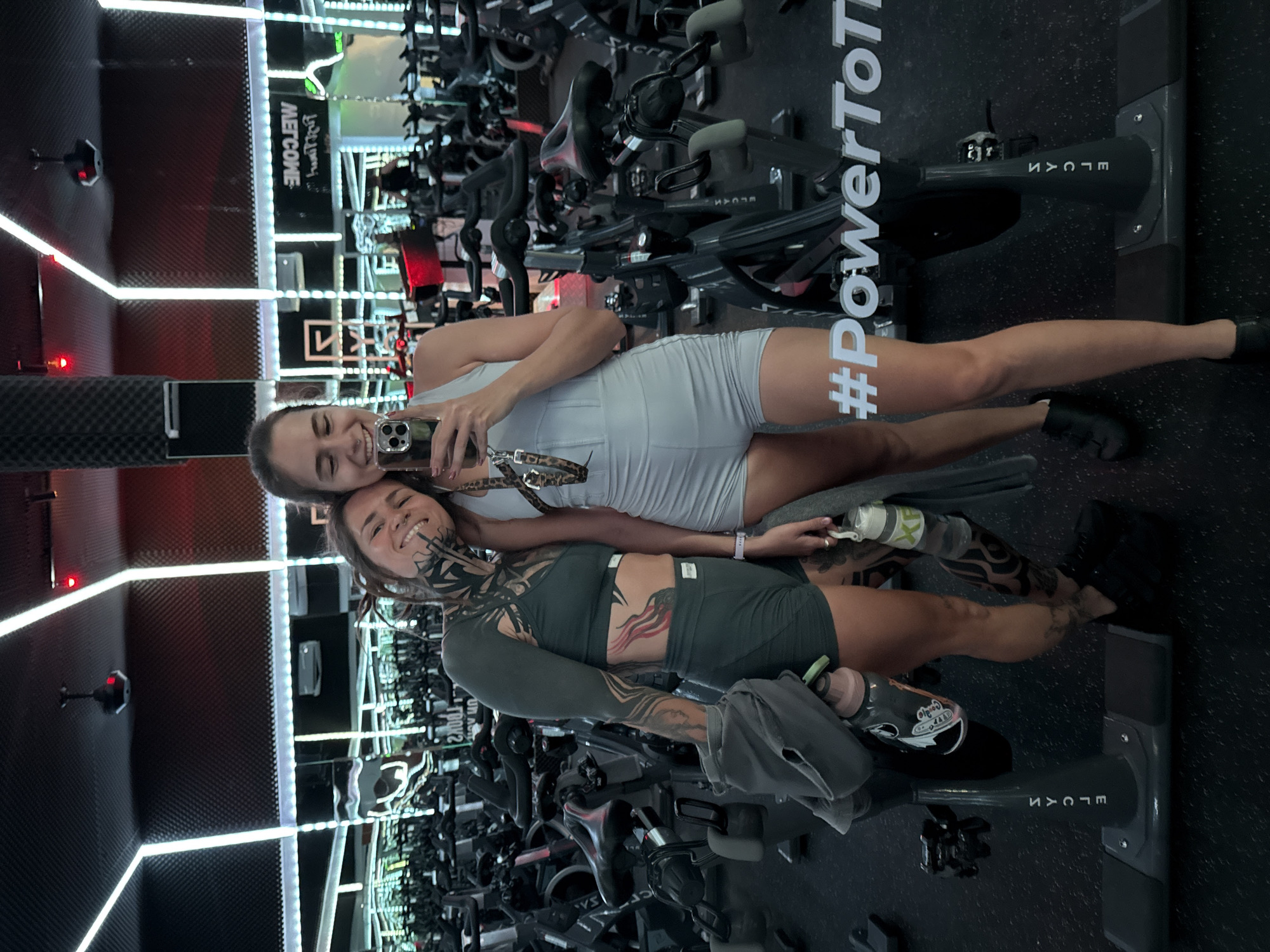 Two women are posing for a selfie in a gym in Canggu, Indonesia. The gym has rows of exercise bikes. The woman on the left has extensive tattoos on her arm, neck, and stomach. She is wearing a dark green athletic outfit. The woman on the right is wearing a light gray athletic outfit and holding a phone. A sign in the background says "WELCOME". The exercise bikes have "ELGAN" written on them. There is a hashtag "PowerToTo" on one of the bikes. The gym has a modern design with neon lights.