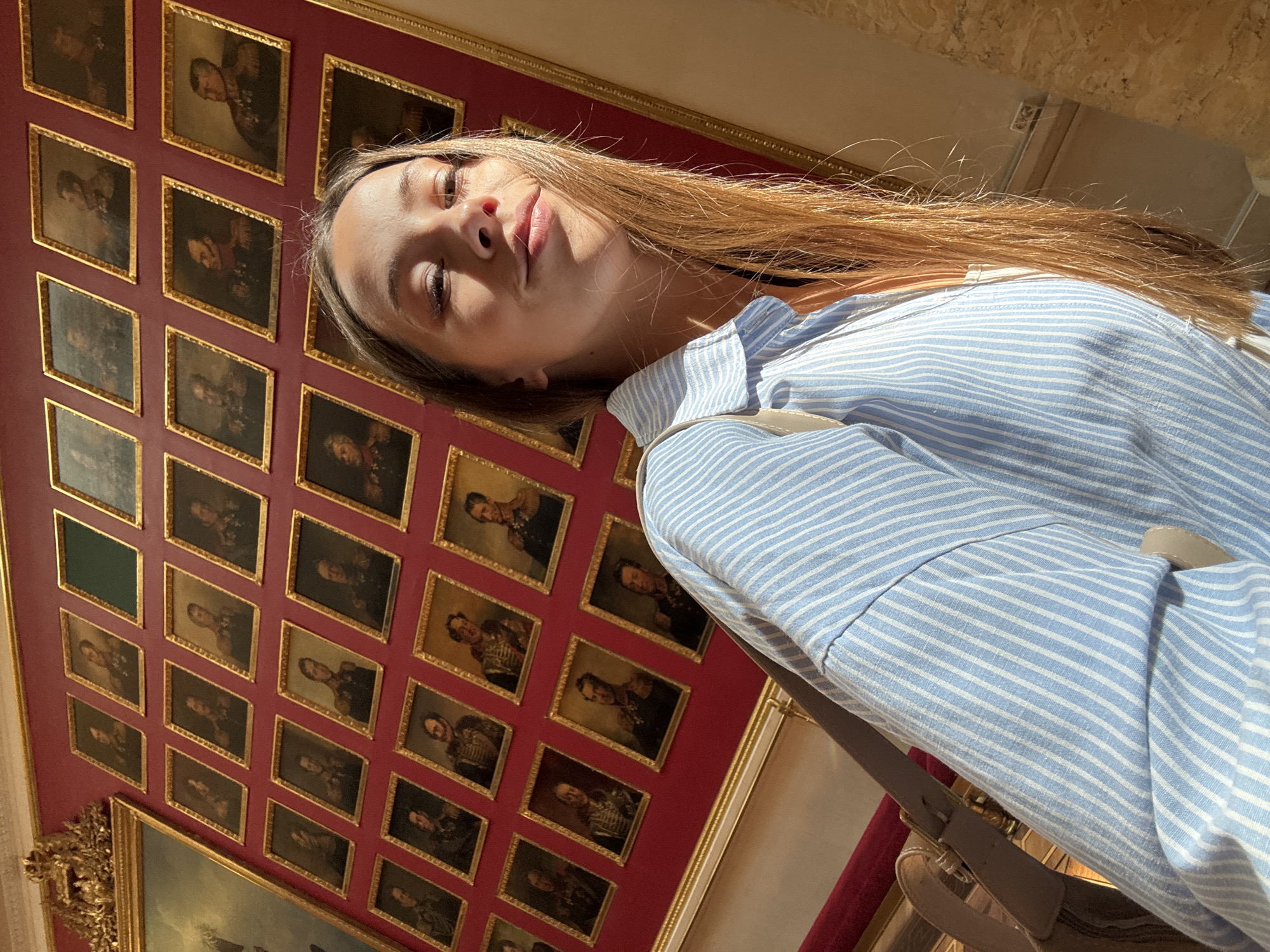 The image shows a young woman taking a selfie in front of a wall of portraits in the State Hermitage Museum, Palace Square, Saint Petersburg, Russia. She is wearing a light blue and white striped button-up shirt and has long, light brown hair. The portraits are framed in gold and appear to be of historical figures, likely military leaders given their uniforms. The wall is a deep red color, and the room has a grand, opulent feel. Sunlight is streaming in from the right side of the image, casting shadows and highlighting the woman's face and hair.