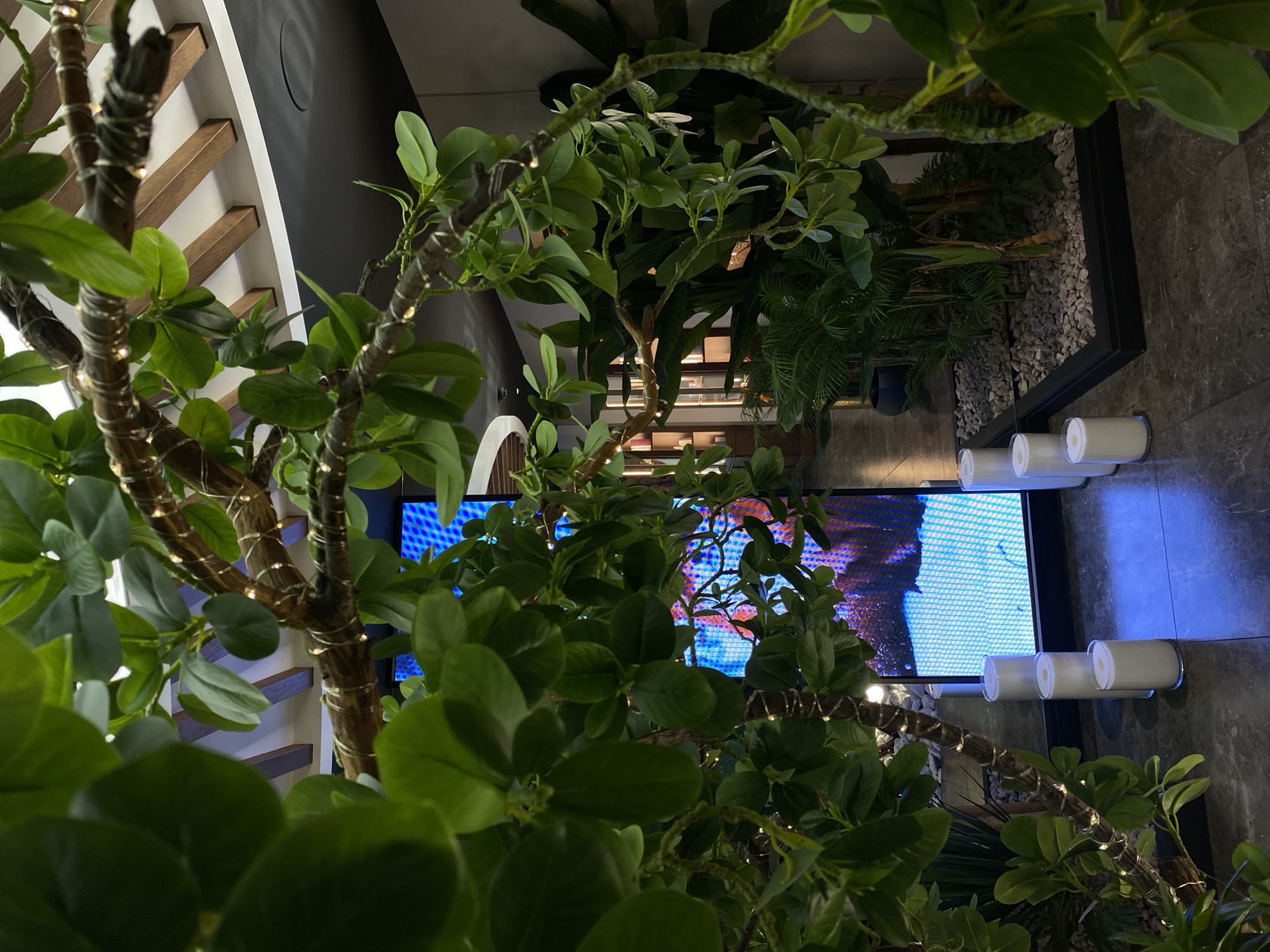 The image depicts an indoor scene, possibly a modern commercial space or a well-decorated residence, with a focus on artificial greenery and ambient lighting. In the foreground, a thick, branch-like structure adorned with small, warm-toned fairy lights winds through the frame, heavily draped with vibrant green artificial leaves. Behind this structure, a large, bright LED screen displays a dynamic image of blue and pink abstract patterns. Several cylindrical, white candles are placed in a row on the dark, polished floor in front of the screen. To the right, a planter filled with grey gravel contains several lush, green artificial plants, including broad-leafed varieties and ferns, creating a dense, tropical aesthetic. The background reveals architectural elements suggestive of a ceiling with wooden beams and a circular light fixture, and further back, a dimly lit display area with shelves filled with objects. The overall impression is one of an artful and curated environment, blending naturalistic elements with contemporary technology and lighting. There are no people visible in the image. The location is not specified as Votkinsk, Russia.