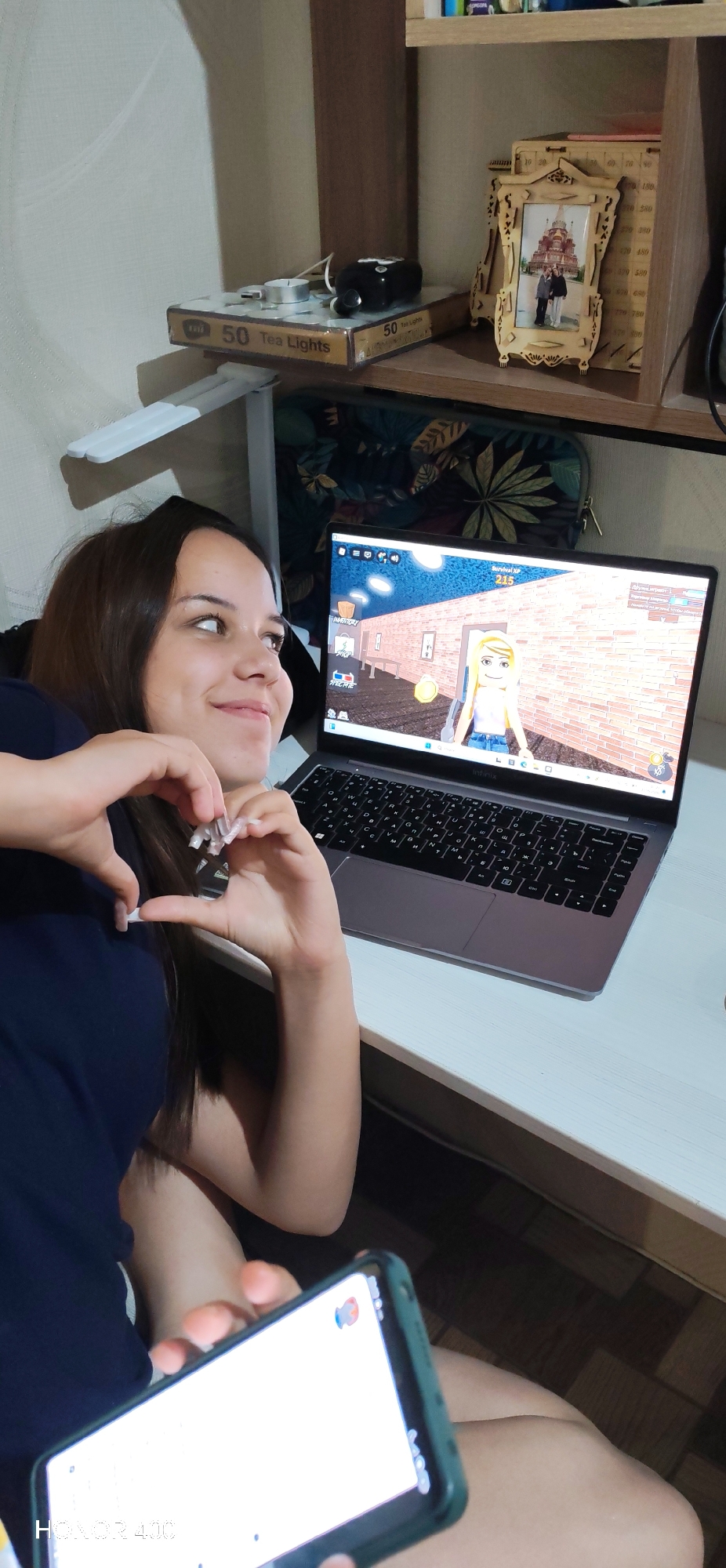 A young woman with dark hair, wearing a dark blue shirt, is sitting at a desk with a laptop and a smartphone. She is making a heart shape with her hands. The laptop screen displays a video game. On the shelf above the desk are boxes of tea lights, a small black device, and a wooden picture frame holding a photo of three people in front of a church. The overall environment appears to be a room in a residence, possibly in Noviy, Russia, given the provided context. The lighting is dim, and the background is cluttered with shelves and a wall.
