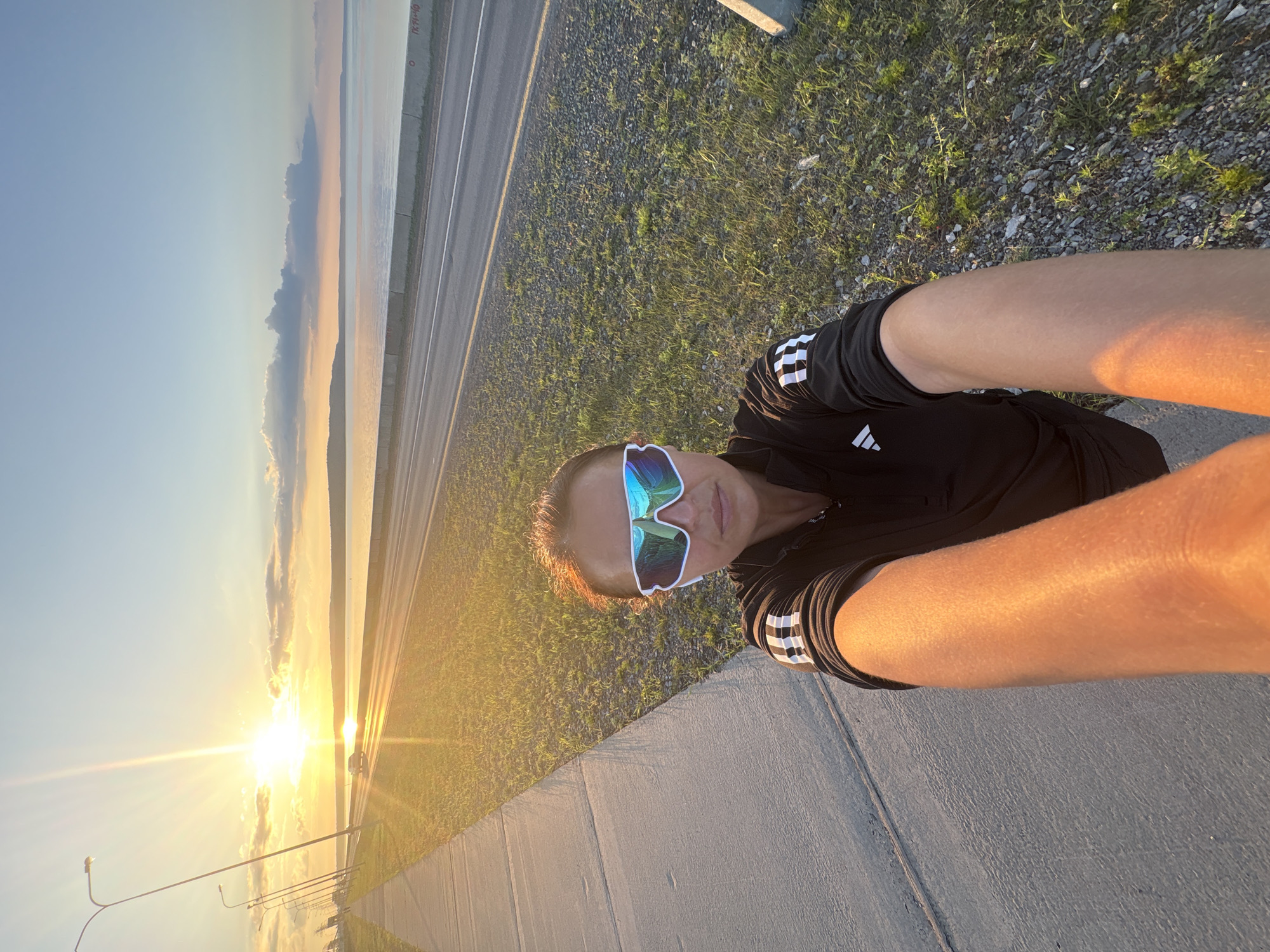 A woman, wearing white-framed athletic sunglasses and a black Adidas jacket with white stripes on the sleeves, takes a selfie at sunrise along a paved walkway next to a road that runs parallel to a large body of water. The sun is low on the horizon, casting a warm, golden light across the scene and creating a lens flare. A car is visible in the distance on the road. The water is calm and reflects the colors of the sky. The walkway is on the left side of the image, bordered by grass and gravel. The location appears to be a bridge or causeway, possibly along the Kama River in Chaikovsky, Russia, given the presence of a road, water, and possibly a bridge structure.
