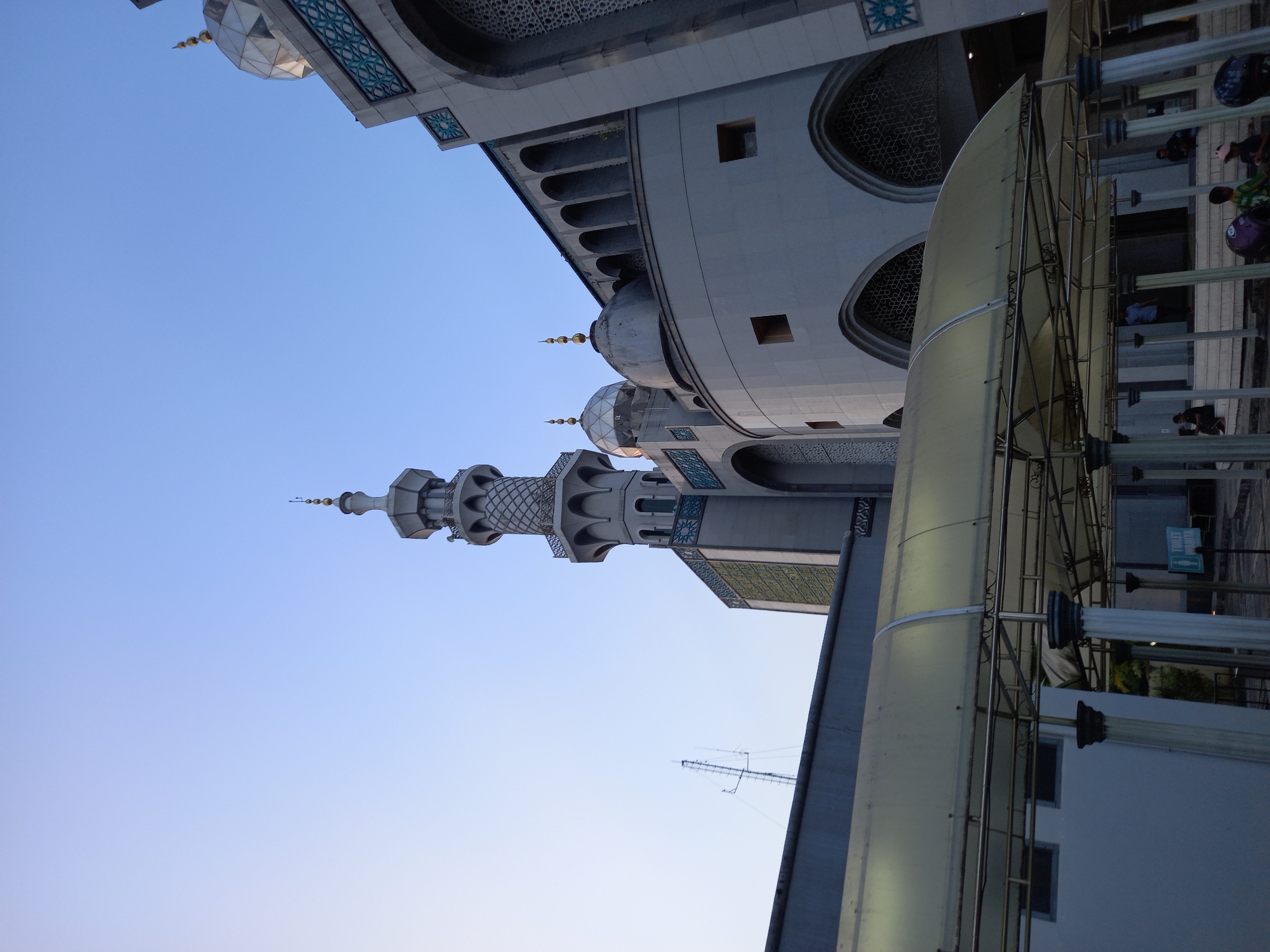 This is a low-angle, exterior, eye-level shot of a modern mosque with traditional architectural elements in Samarinda, Indonesia. The sky is clear and pale blue, suggesting it might be early morning or late afternoon. The main subject is the mosque's structure, which is predominantly light gray and features multiple domes and a tall, slender minaret. The minaret has an intricate, latticed design and tapers towards its finial. The mosque's facade includes arched windows with decorative geometric patterns and rows of smaller arched openings. In the foreground, a covered walkway with a translucent yellow roof and metal supports runs horizontally across the frame, obscuring some of the lower part of the mosque. Several people are visible in the background, some seated and some standing under the walkway or near the mosque entrance. A "TOILET WANITA" (Women's Toilet) sign is visible. A tall antenna or communication tower stands to the left of the minaret. The overall impression is one of a large, active religious site.