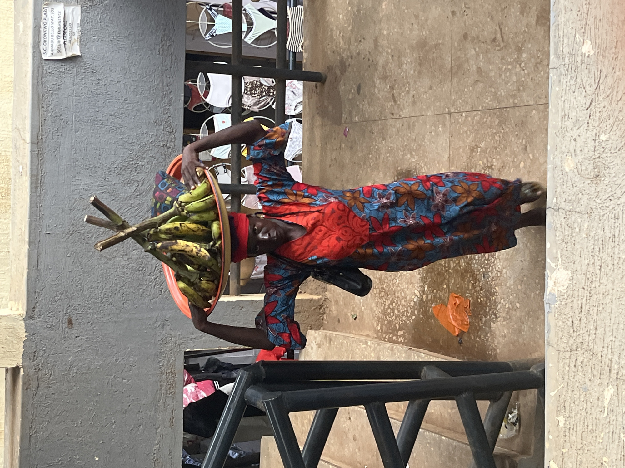 A woman carries a large tray of bananas on her head, walking down a concrete stairway. She wears a brightly colored, floral patterned dress and a red headscarf. A black bag is slung over her shoulder. Behind her, a stall displays various undergarments for sale, and a sign on the wall reads "S.C. OKONKWO PLAZA AHMADU BELLO WAY, JOS SECURITY EMERGENCE PLEASE CALL 08165927149 OR 07039374700". The scene appears to be in an outdoor market or public space in Jos, Nigeria.