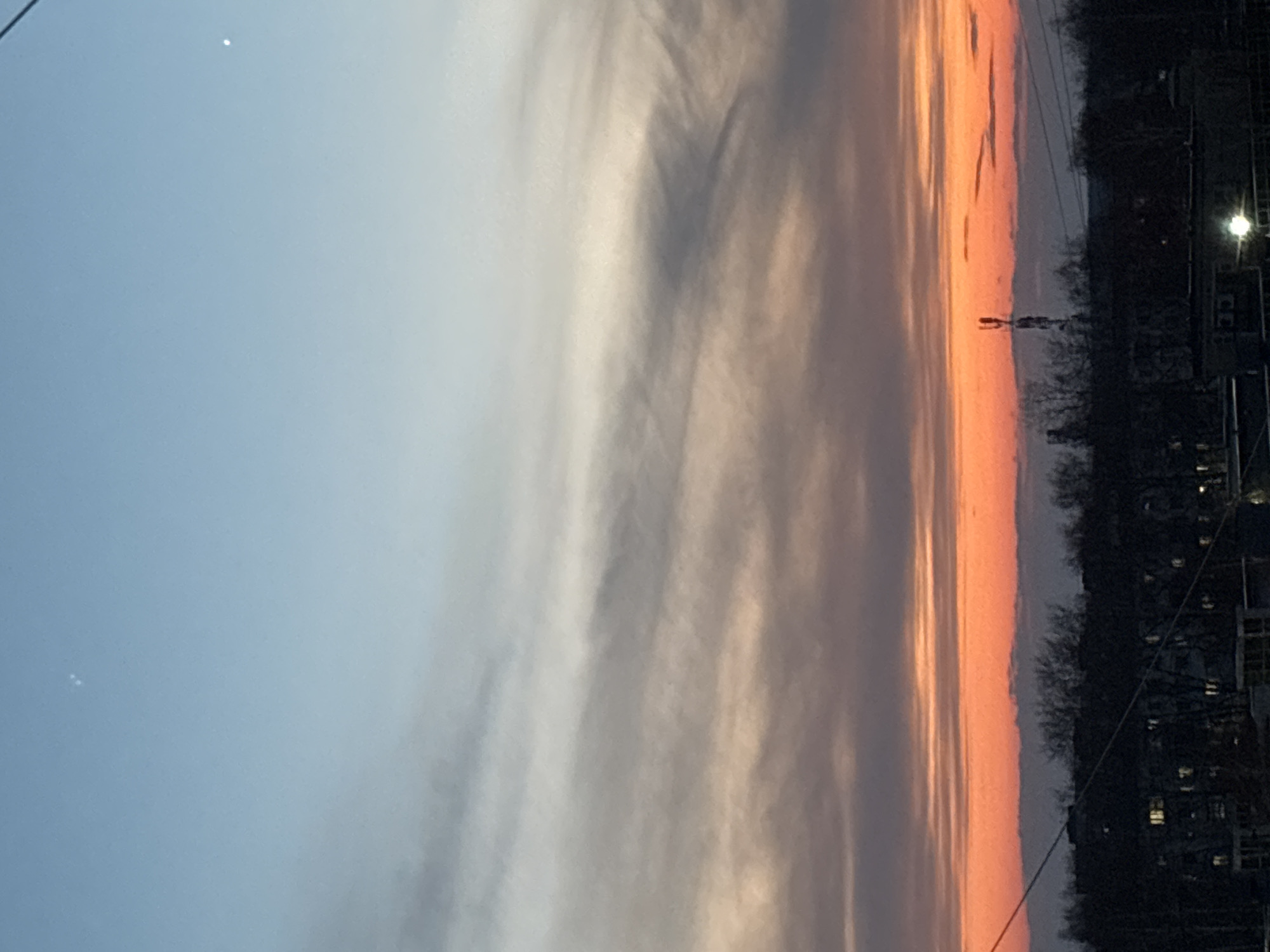 The image shows a sunset in what appears to be a city environment, likely in Chaikovsky, Russia given the prompt. The sky transitions from a pale blue to an orange-red near the horizon, with streaks of white and grey clouds. Silhouettes of trees and buildings are visible along the horizon line. A streetlight illuminates the scene, indicating it is dusk or early evening.