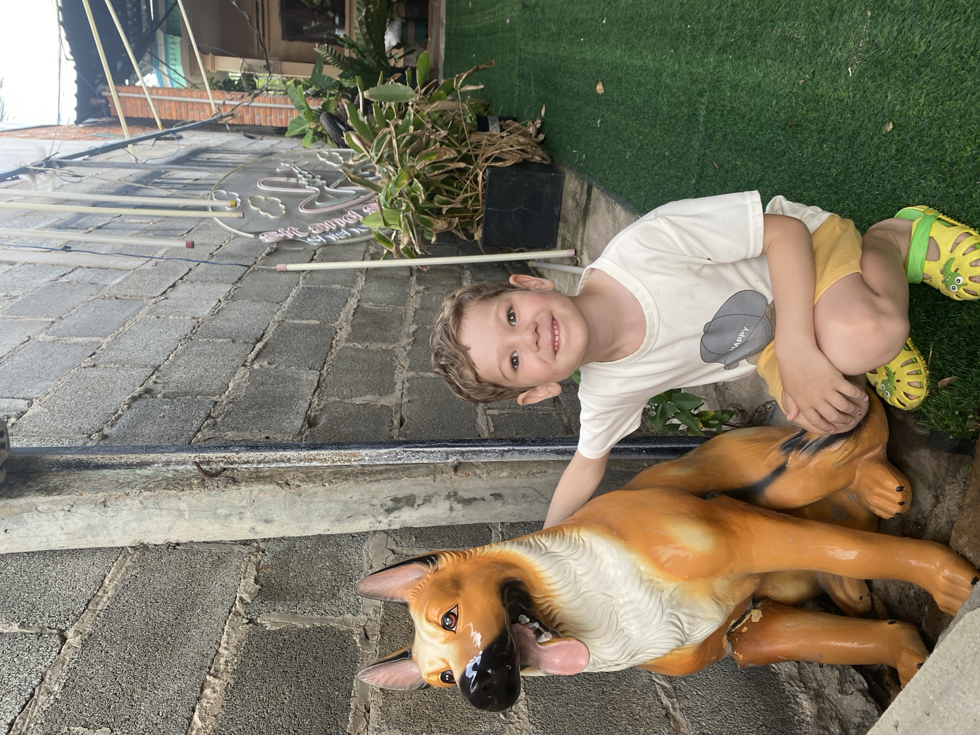 A young boy kneels beside a painted German Shepherd statue outdoors in Phu Thuy Ward, Vietnam. The boy wears a white t-shirt, yellow shorts, and green/yellow Crocs. The statue is lying on its side on the gray brick-paved ground. Behind them is a gray brick wall and some greenery near artificial turf. There is a neon sign on the wall behind the bricks.