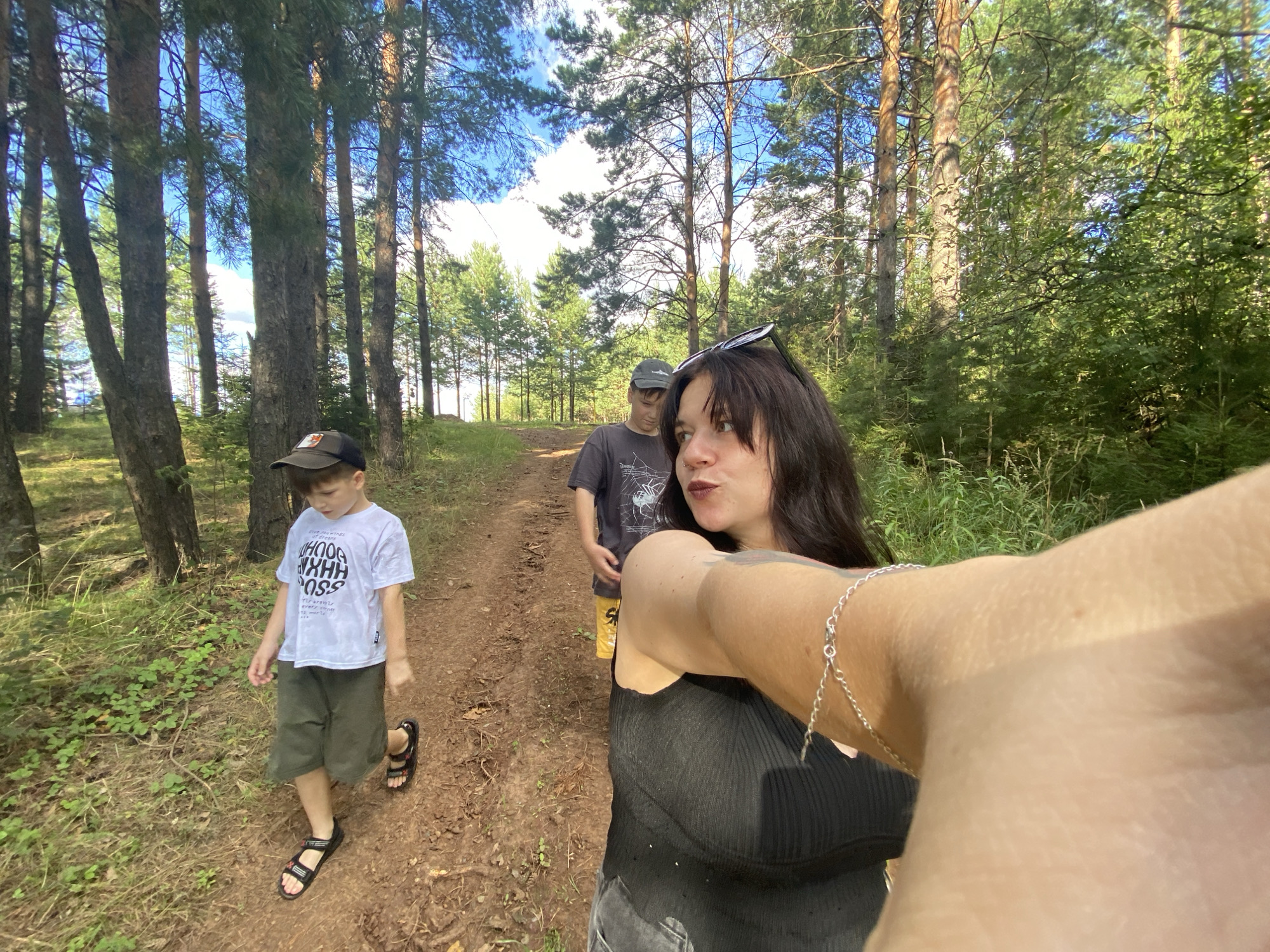 A selfie is taken by a young woman with dark hair and sunglasses perched on her head, wearing a black tank top and a silver bracelet. She is in the foreground, looking to the right. Behind her, on a dirt path in a forest, a young boy in a white t-shirt and khaki shorts, wearing a black baseball cap, walks ahead. Further behind him, a person in a grey t-shirt and yellow shorts is walking. The environment is a forest with tall pine trees and green foliage under a bright blue sky with some clouds. The path appears to be in a rural or natural setting, possibly on the outskirts of the city of Фоки, Russia.