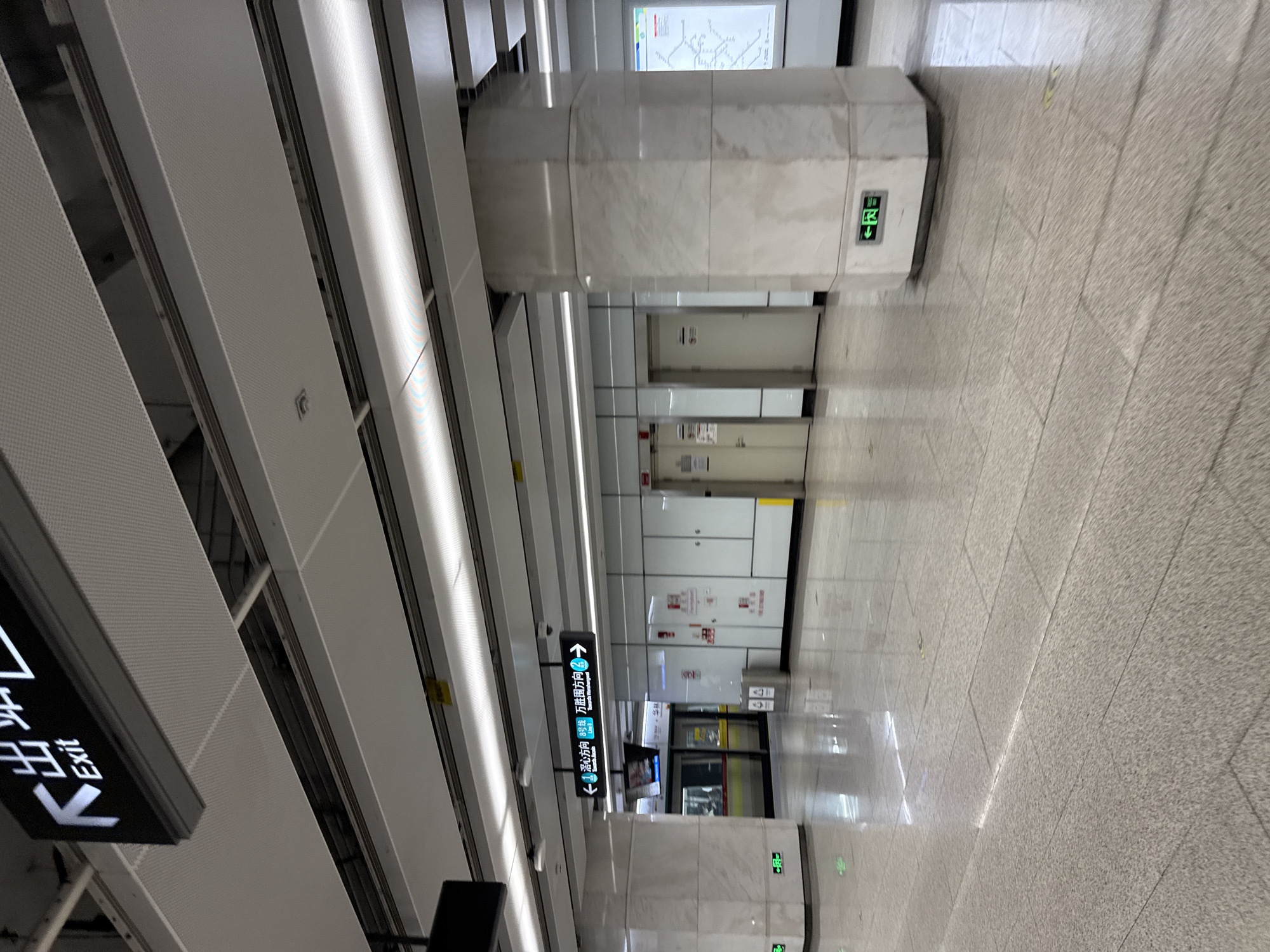 The image shows an interior view of a modern subway station in Baiyun District, China. The station features a clean, brightly lit environment with polished marble floors and white, paneled walls. A prominent overhead sign with Chinese characters and the English word "Exit" is visible in the upper left corner, indicating an exit route. Below this, another sign with directional arrows in Chinese and English points towards "Xiangyun North Station" and "Wanshangyuan Station".
In the background, a subway train is visible at the platform, with its doors open. Several doors, presumably to other areas of the station or maintenance rooms, are present along the walls, some with emergency signage. A large, octagonal marble-clad pillar stands in the mid-right of the frame, with a green illuminated exit sign attached to its base. Another similar green exit sign is seen on the floor in the mid-left. The overall impression is one of a functional and well-maintained public transportation hub. There are no people clearly visible in the image, suggesting it was taken during a quiet period or from a distance where individuals are not distinct.