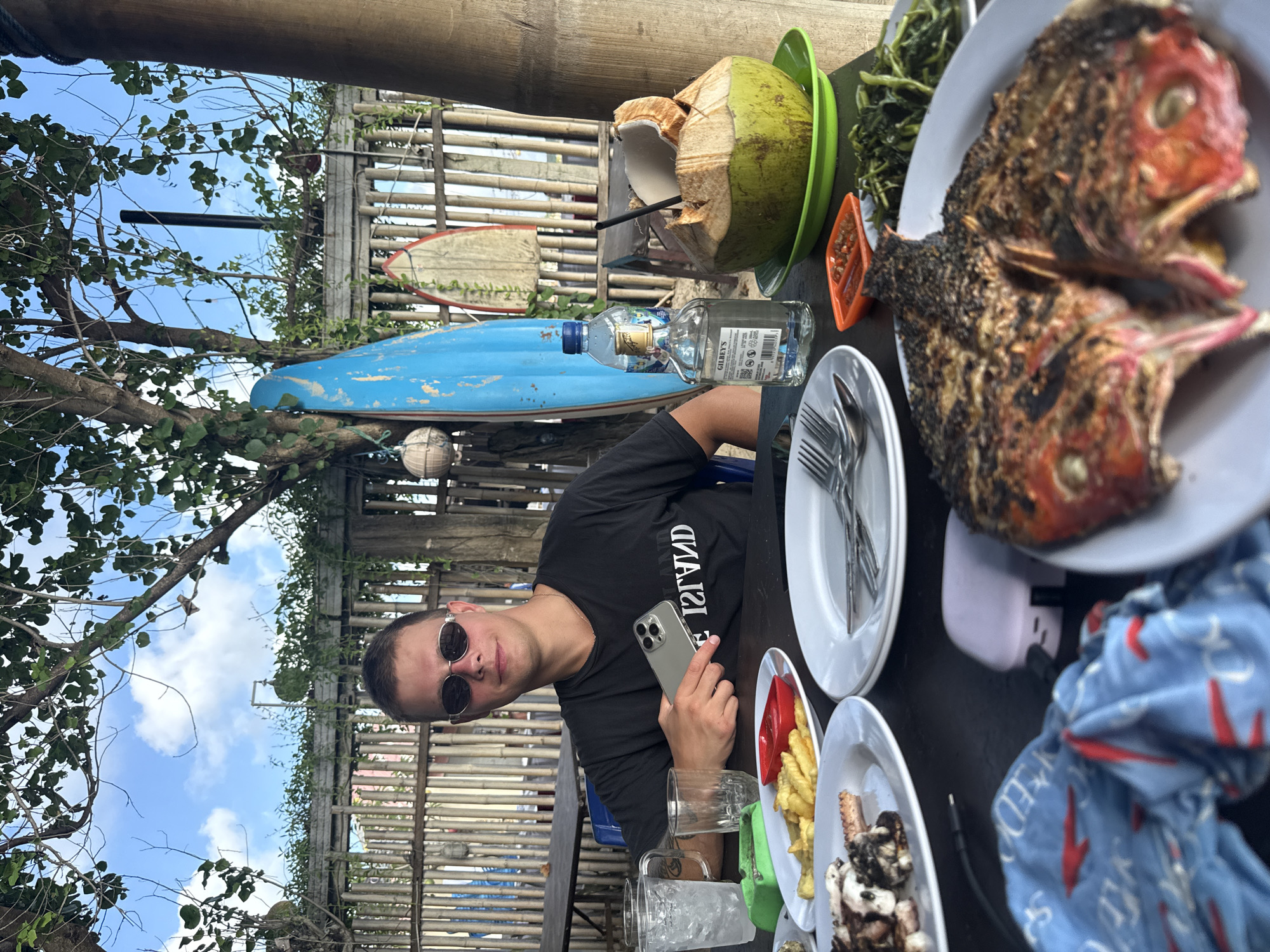 A man with short brown hair and sunglasses sits at a dark wooden table in an outdoor setting. He is holding a silver-colored smartphone in his right hand. The table is laden with plates of food, including a grilled fish, fries, and other dishes, along with a coconut drink and a bottle of Gilbey's gin. In the background, there are bamboo structures, surfboards, and trees against a bright sky. The location is likely Kuta, Indonesia, judging from the tropical environment and surf culture elements.