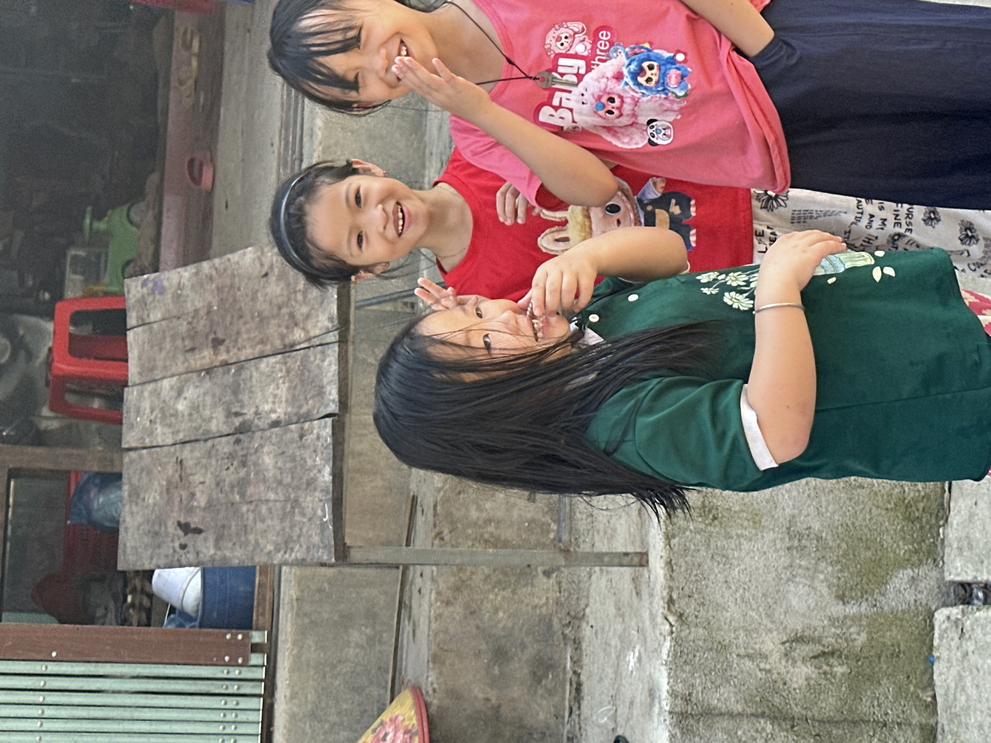 Three girls are positioned closely together outside in Pờ Ly Ngài Commune, Vietnam. The topmost girl wears a pink shirt with cartoon characters, dark pants, and has short dark hair. The middle girl wears a red shirt and a black headband. The bottom girl wears a green shirt and has long dark hair. All three girls are smiling. A weathered wooden table is to the left. A red plastic chair and other objects are visible in the background. The ground and a low wall are concrete.