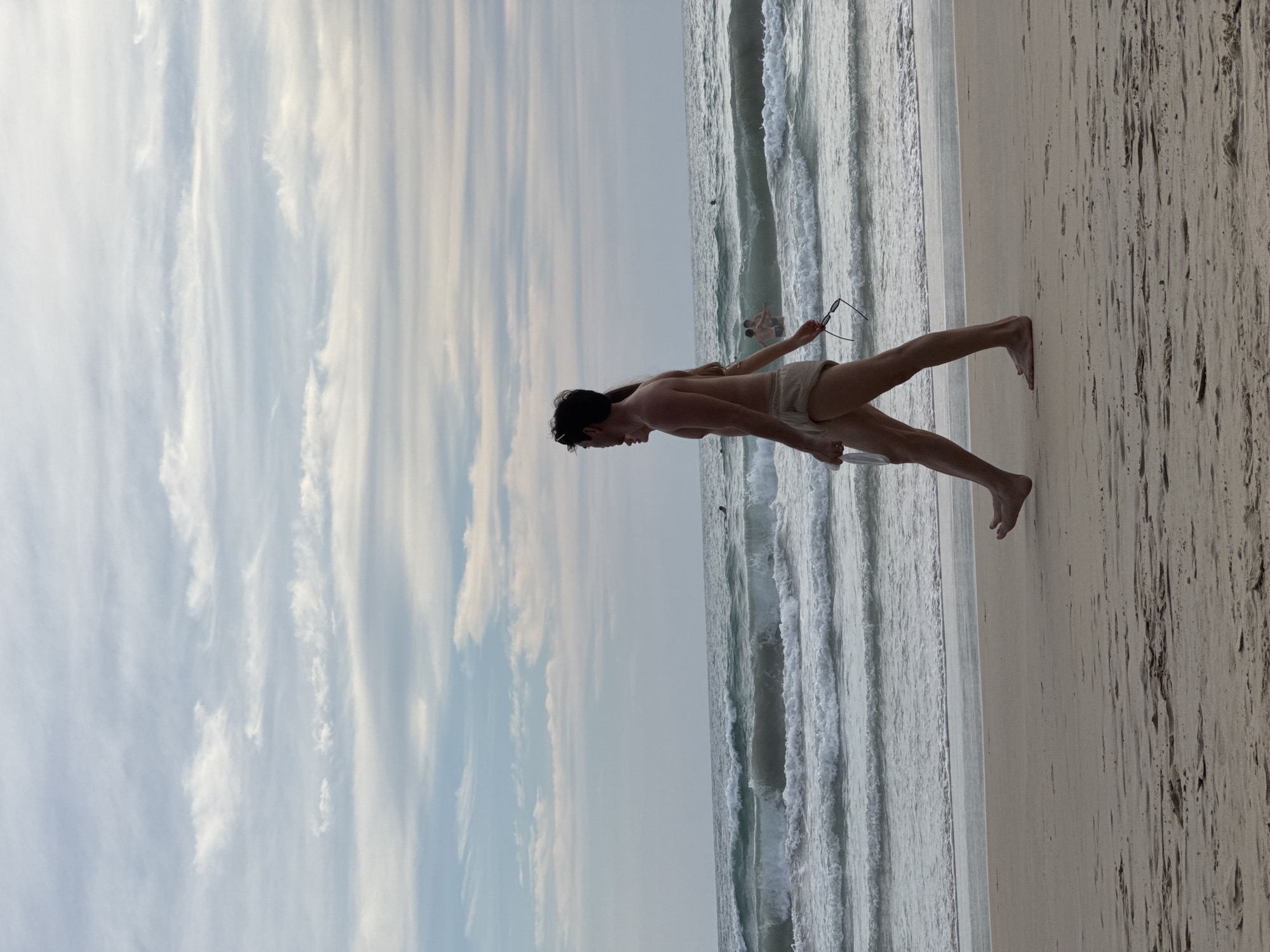In Karon, Thailand, on a sandy beach, a couple walks along the shoreline under a cloudy sky. The man, in focus, is in the foreground, wearing light-colored swim shorts. He is holding a white object in his left hand and a pair of dark sunglasses in his right. A woman, partially obscured by the man, is walking beside him. In the background, the ocean displays waves with whitecaps, and several people are visible in the water. The sand is a pale beige, showing footprints, and the water is a muted greenish-blue. The sky is filled with a mix of white and grey clouds, streaked across a pale blue canvas.