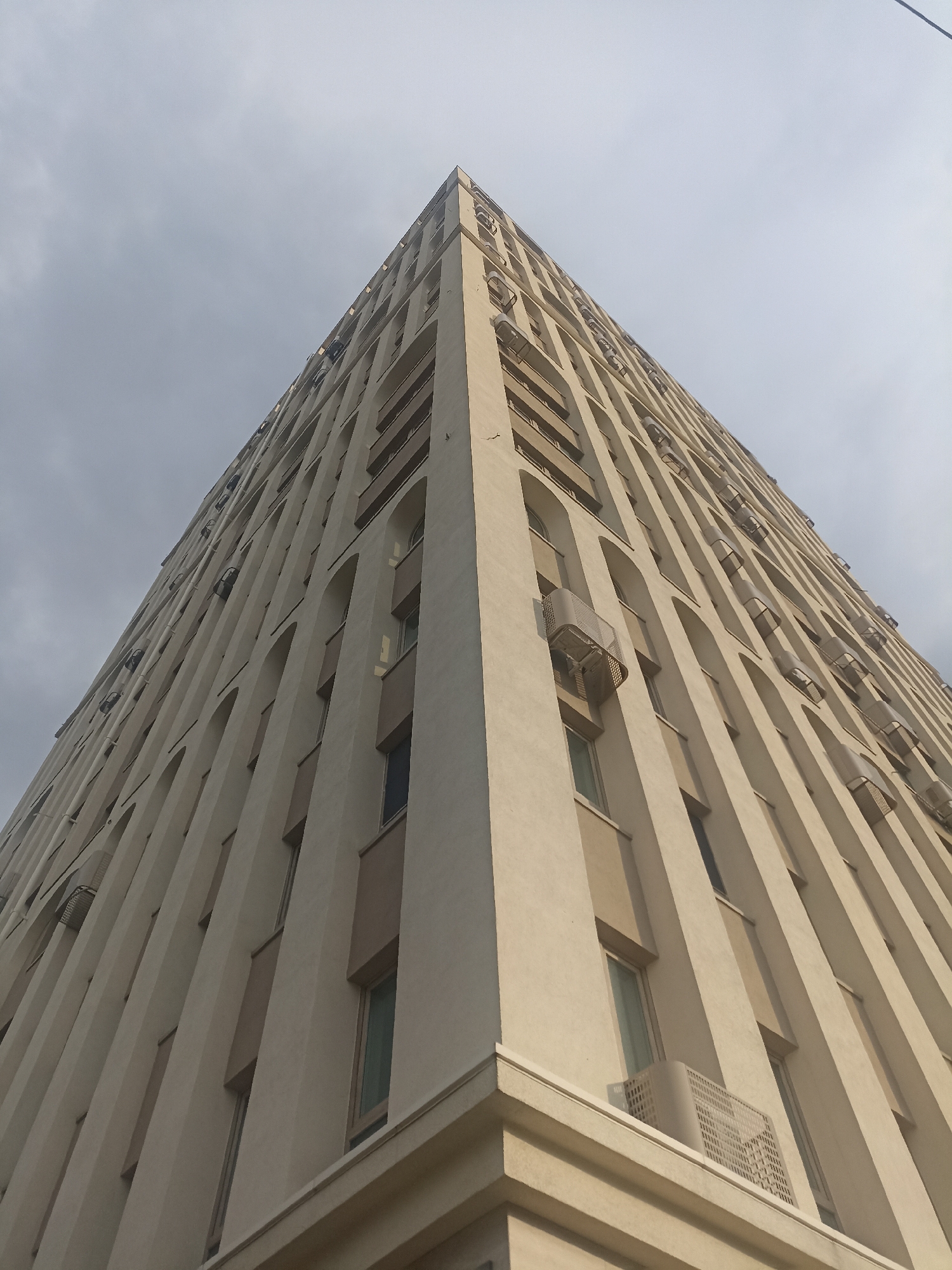 The image shows a low-angle, wide shot of a tall, beige apartment building with a modern architectural design. The building has a prominent corner that extends upwards, creating a dramatic vanishing point. Numerous windows are visible, many with arched tops, and some are partially obscured by horizontal balconies. Several outdoor air conditioning units are mounted on the building's facade, some with visible grates. The sky above is overcast and appears to be a light gray color. The building is positioned in a way that suggests it is a significant structure within its urban environment. The metadata indicates the image was taken in Novosibirsk, Russia, on March 19, 2023. There are no people visible in the image.