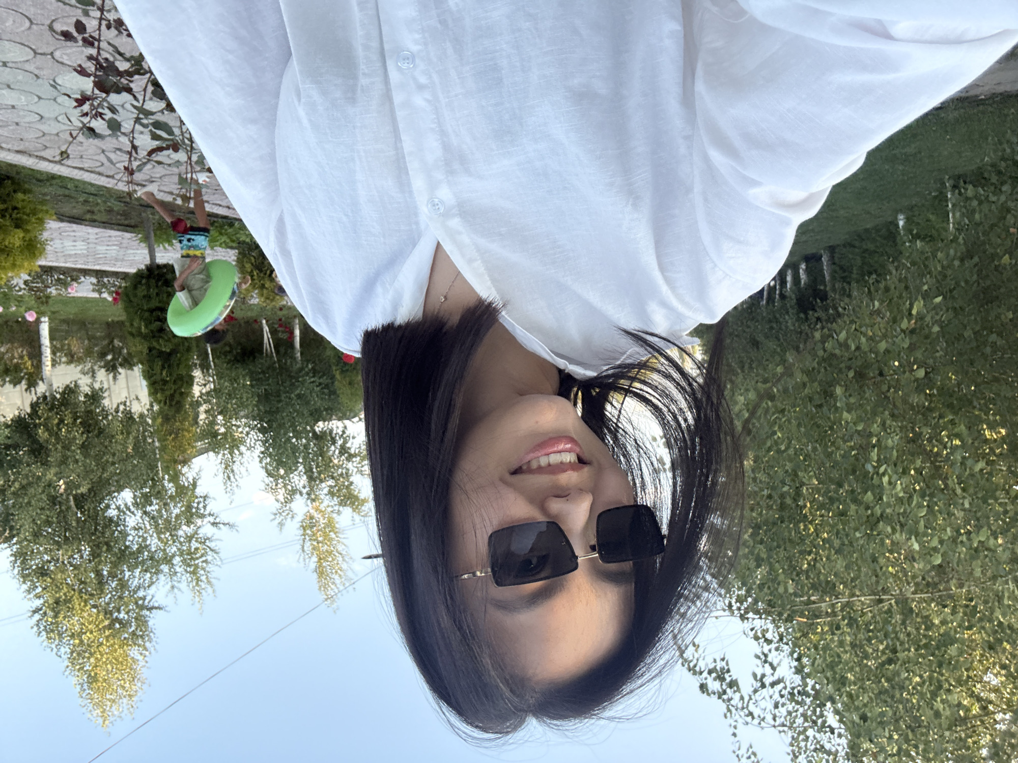 A young woman with dark hair and sunglasses is taking a selfie outdoors. She is wearing a white collared shirt. Behind her, a boy in a green shirt and patterned shorts walks away, carrying a green inflatable ring. The background shows trees, a pathway, and what appears to be a residential area, suggesting a location in Sary-Oy, Kyrgyzstan. The lighting indicates it is daytime, likely late afternoon due to the warm tones.