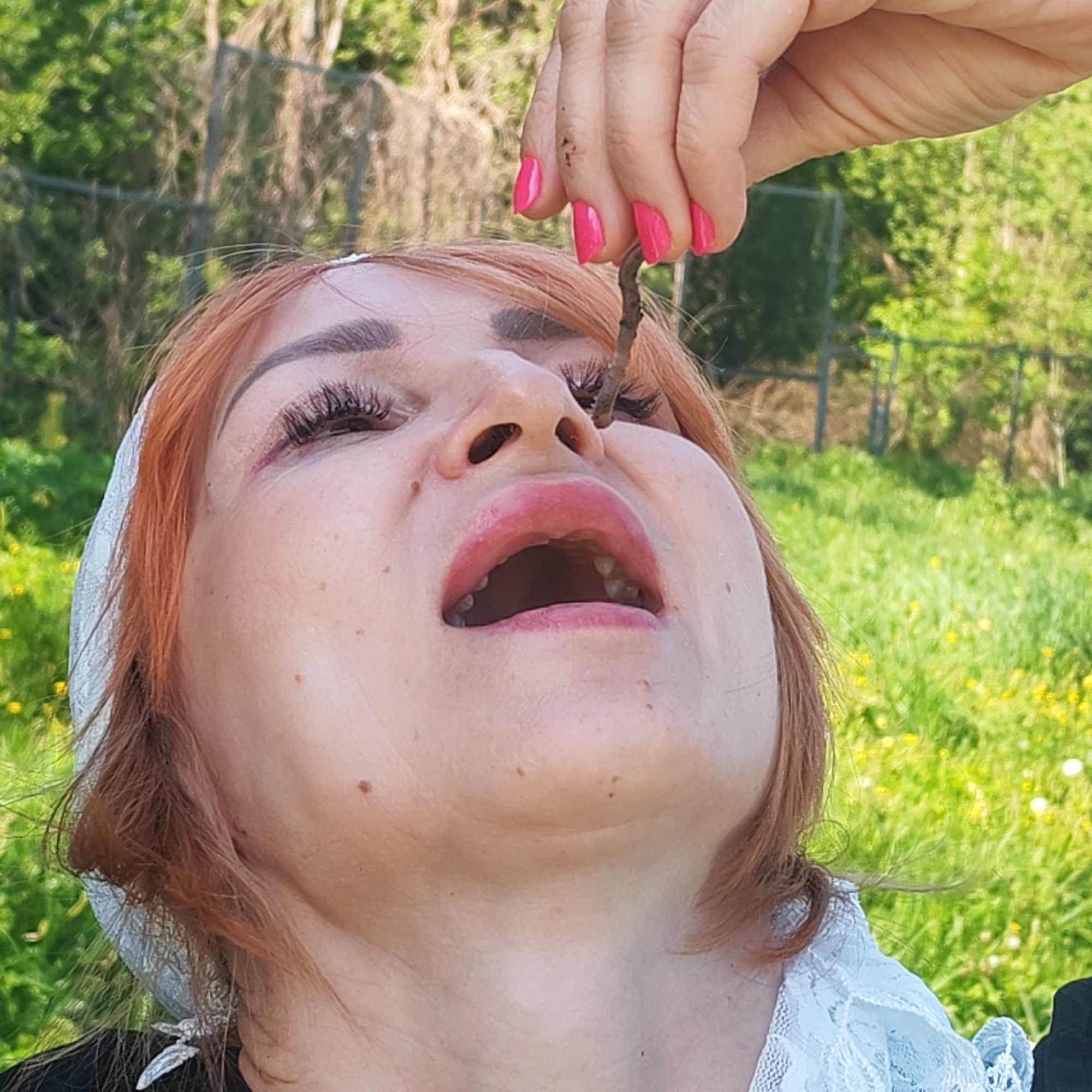 A woman with fair skin, red hair, and pink lipstick is positioned in the foreground. Her head is tilted back, mouth open, and a hand with pink nail polish is holding a small twig near her nose. The background includes green grass with scattered yellow flowers and blurred trees. The image does not provide sufficient information to confirm the location in Malyi Bukor, Russia.