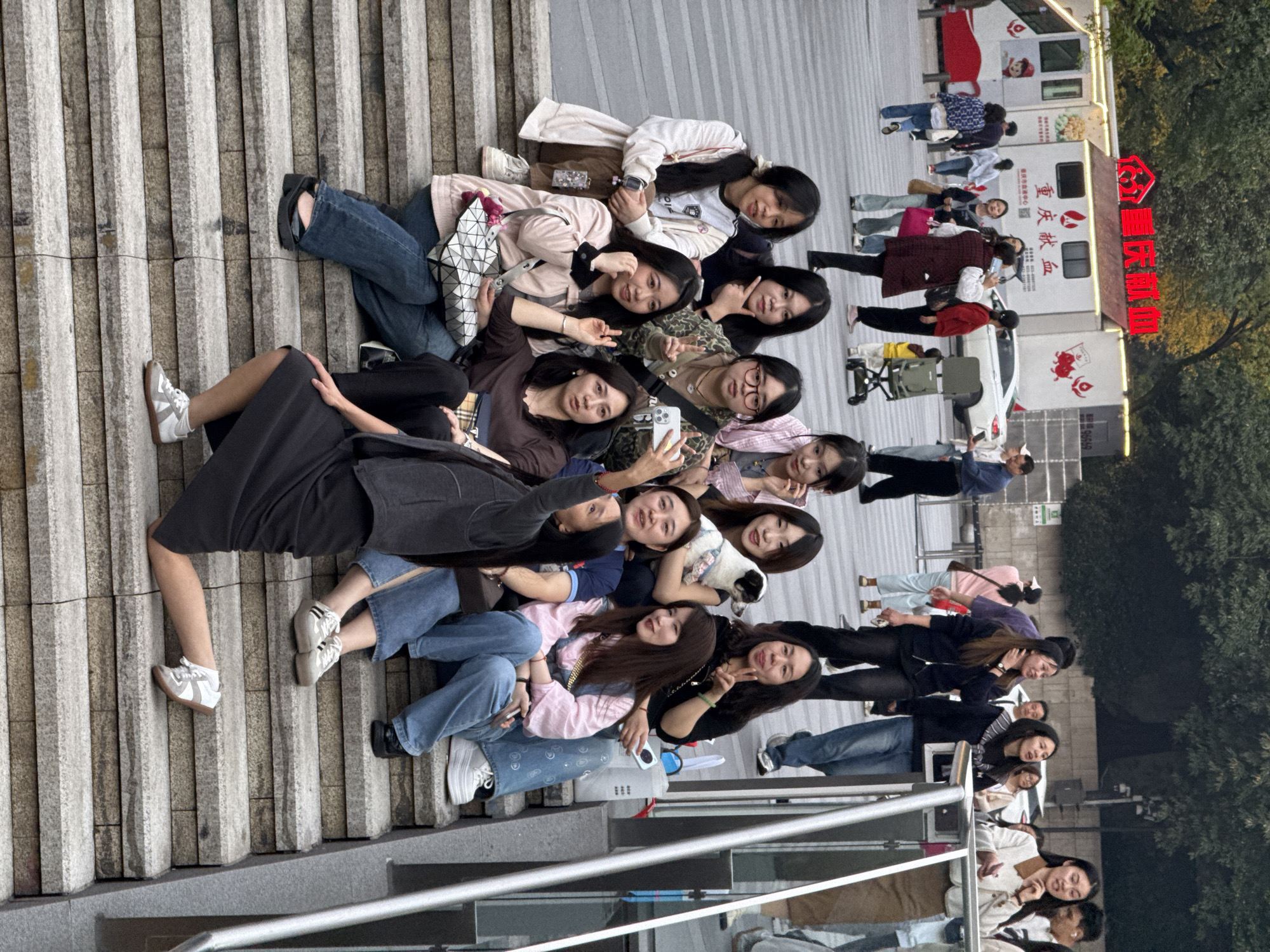 A medium-angle, outdoor shot shows approximately 14 young adult women sitting on concrete steps in Yuzhong District, China. They are lined up on the steps facing forward and are posed for a group photo. Some are holding up peace signs. One woman in the middle is holding a phone, presumably taking the photo. They wear a variety of casual clothing, including jeans, dresses, blouses, and jackets.
In the background, a white van and other vehicles are visible, as well as pedestrians and some trees. Red Chinese characters are prominently displayed on a nearby building or structure. The sky appears overcast. The steps are light gray, textured concrete. A railing is visible in the foreground.