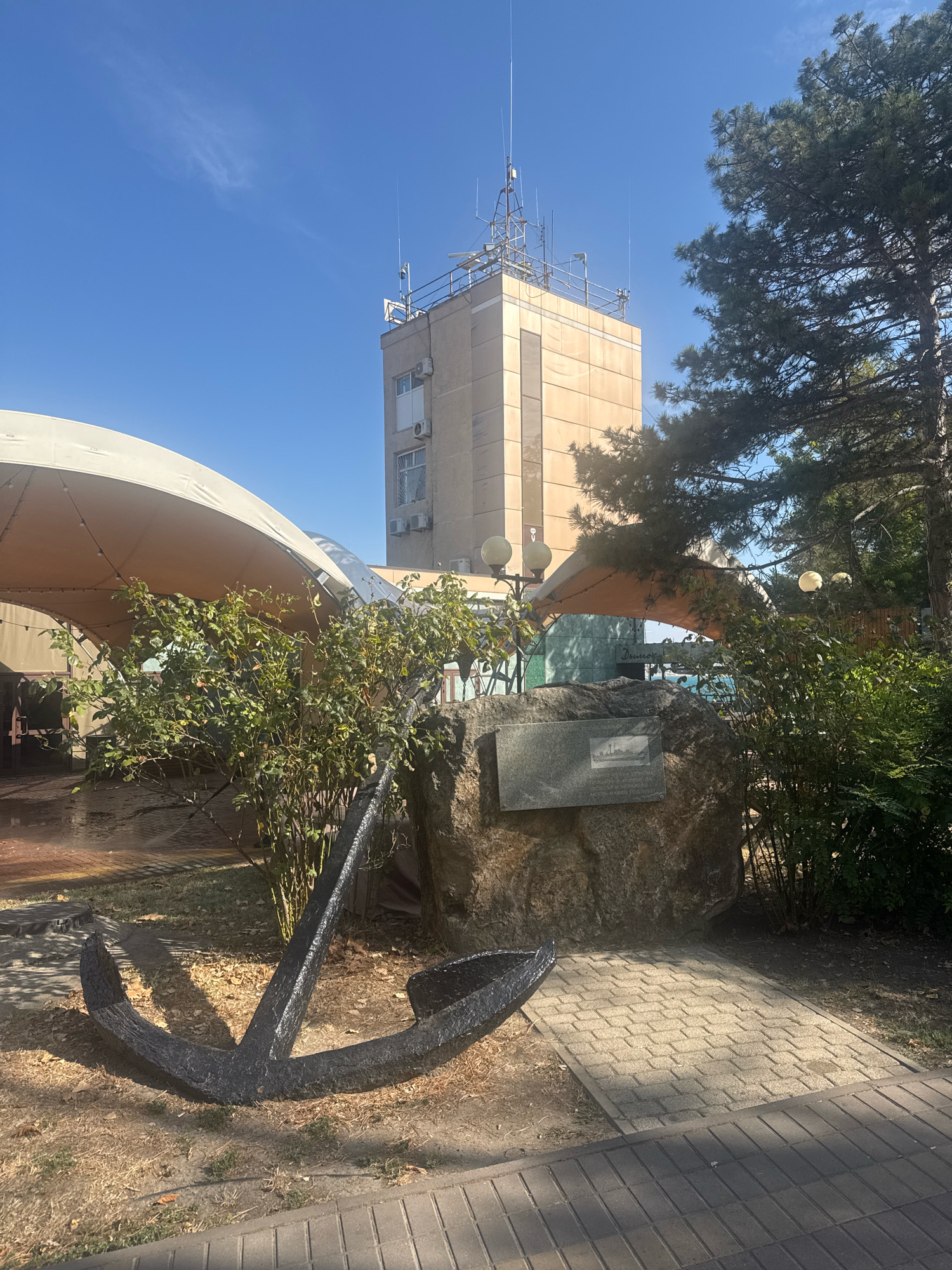 An exterior view shows a tall, beige building with antennas on top, situated next to a large, dark anchor resting on dry grass and dirt. The anchor is positioned in the foreground, to the left of a large rock with a plaque affixed to it. Behind the anchor and rock, a large, light-colored awning covers an outdoor seating area. Lush green bushes are visible around the rock and extending towards the right side of the image. A paved walkway runs along the bottom right, leading towards the anchor and rock. The sky above is a clear blue with a few wispy clouds. This scene is located at the Anapa Regional Museum in Anapa, Russia, which focuses on the history of the city and the Black Sea.