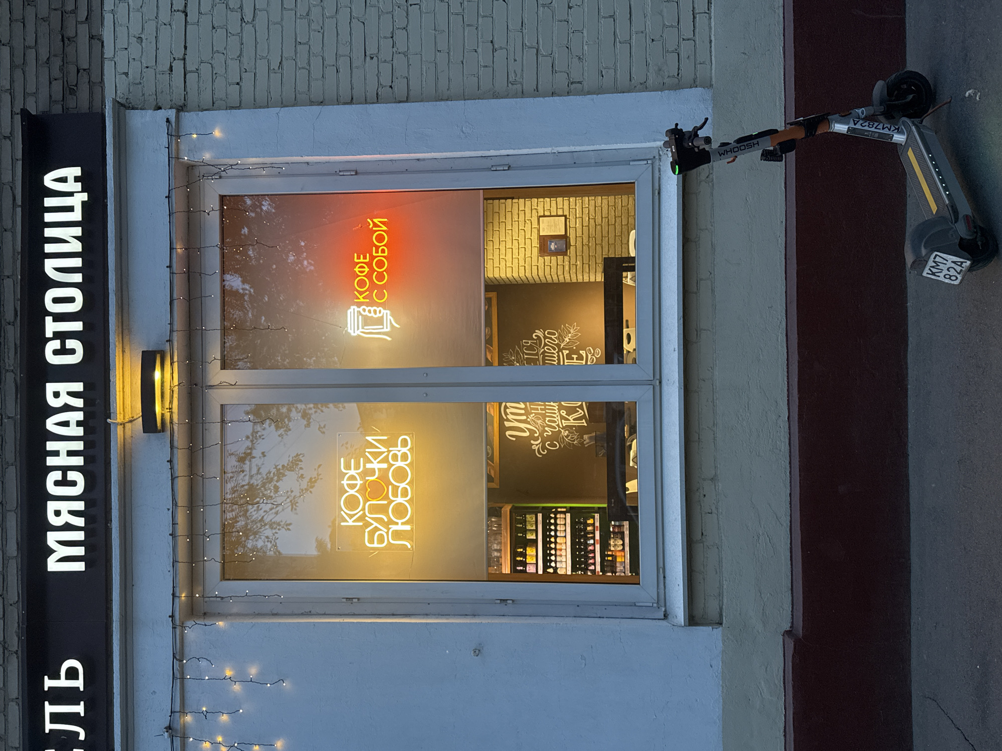 An outdoor, street-level view shows the illuminated facade of a commercial establishment, likely a cafe or shop, at dusk or night. The scene is brightly lit by artificial lights against a dim background.
On the left, a tall, vertical black sign with white illuminated Cyrillic letters reads "МЯСНАЯ СТОЛИЦА" (Myasnaya Stolitsa, meaning "Meat Capital"), with the letters "ЛЬ" partially visible at the bottom, suggesting a longer word or another sign beneath. The building wall behind the sign is made of light-colored brick.
The central focus is a large window, framed in white, decorated with delicate string lights. Inside the window, several neon signs are visible. A red neon sign in the upper section reads "КОФЕ С СОБОЙ" (Kofe s Soboy), which translates to "Coffee to Go" or "Coffee with You," featuring a small icon of a coffee cup. Below, a yellow neon sign proclaims "КОФЕ БУЛОЧКИ ЛЮБОВЬ" (Kofe Bulochki Lyubov), meaning "Coffee, Buns, Love." Through the window, the interior shows a brick wall, shelves stocked with various items, and a dark wall with white chalk-style text, partially visible as "УТРО с чаши..." (Utro s chashi...), likely part of a phrase about morning coffee.
To the right of the window, an electric scooter is parked upright on the pavement. The scooter is silver and black, with the brand "WHOOSH" visible on its stem. A license plate or identification tag on the scooter's rear fender and top stem both read "KM7 82A" and "KM782A" respectively. The surrounding building wall transitions from a light beige to a darker red-brown. The pavement surface is dark gray asphalt.
The overall impression is that of a welcoming, active business operating in an urban setting in Moscow, Russia.