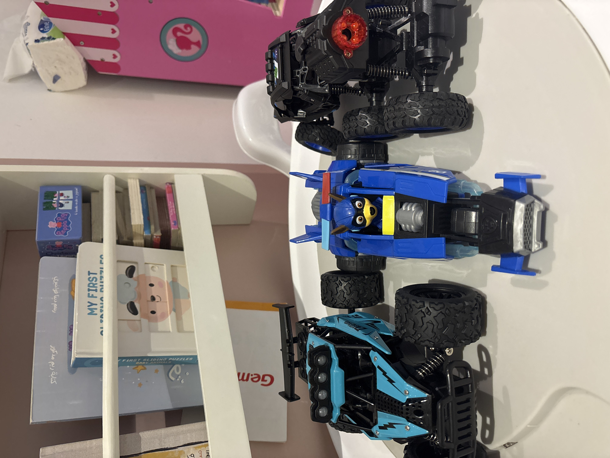 Three remote-controlled toy cars are displayed on a white surface, possibly a tray or table. From left to right, the first car is blue with black accents, featuring a spoiler and off-road tires. The second car is bright blue and appears to be designed to resemble a character, possibly a superhero, with a yellow face visible in the cockpit. The third car is black and also has off-road tires and a spoiler, with a red taillight. Behind the cars, a shelf holds several children's books. A pink structure with vertical stripes and a circular graphic of a silhouette is visible on the right side of the image. A partially visible roll of tissues is perched on top of the pink structure. The background is a light pink wall. There is no visible indication of the city of Dubai, United Arab Emirates.