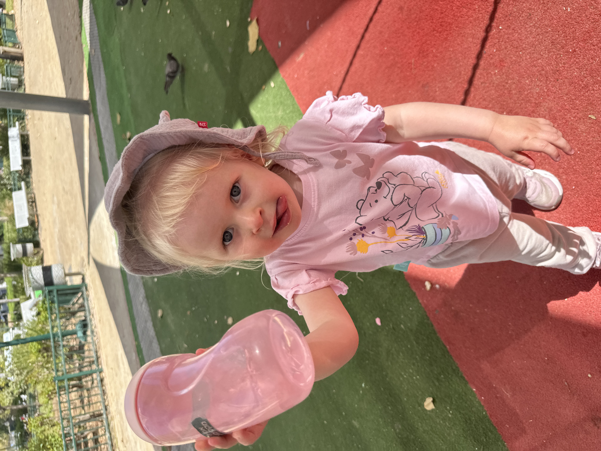 This brightly lit, vertically oriented image captures a young child on an outdoor playground in Petah Tikva, Israel. The blonde-haired, blue-eyed child, appearing to be a toddler, is centered in the frame, looking up at the camera with a playful expression, their tongue slightly extended. They are wearing a light pink short-sleeved t-shirt with a graphic featuring a bear (resembling Winnie the Pooh) and colorful floral designs, white pants, and light pink and white sneakers. A light brown sun hat, with a small red "m.cc" tag visible on its side, covers their head.
In the child's left hand, they hold a transparent pink plastic bottle with a white lid, which has the text "DDN PPD" visible in white letters. The ground beneath the child is a safety surface, divided into vibrant red and green sections. Bright sunlight casts distinct shadows on the red portion of the ground, suggesting a clear, sunny day, likely during the morning or early afternoon.
In the background, several pigeons are scattered across the green playground surface. Beyond the immediate play area, there's a concrete path, followed by lush green foliage, trees, and a metal fence or railing. A silver pole, possibly part of playground equipment or a structural support, is visible on the far left. The overall scene conveys a cheerful and active moment of a child enjoying the outdoors.