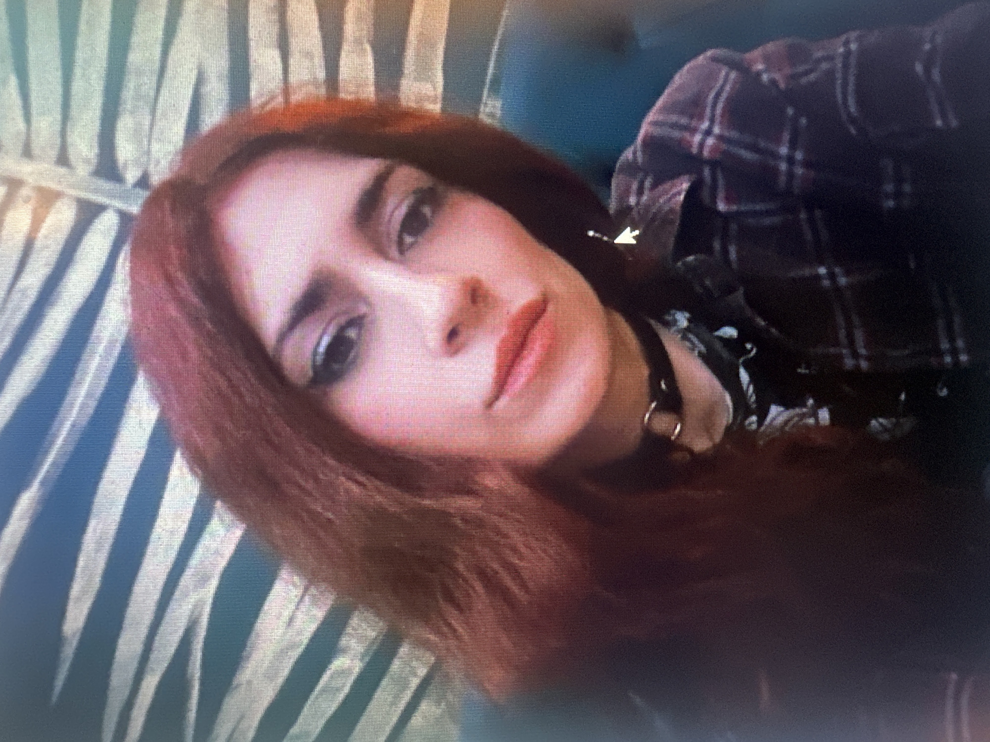 A medium shot photograph shows a woman with long, auburn hair. She is wearing a dark choker necklace, a black and red plaid shirt, and a dark, patterned shirt underneath. Her makeup includes winged eyeliner and red lipstick. She is looking towards the camera with a slight smile. The background consists of a textured wall with blue and white stripes. The photograph appears to be taken indoors, possibly in a home or a dimly lit establishment. The overall lighting is soft, with some graininess visible, suggesting it may have been taken with a low-quality camera or under low-light conditions. There is no discernible information within the image to suggest a specific location in the city of Togliatti, Russia.