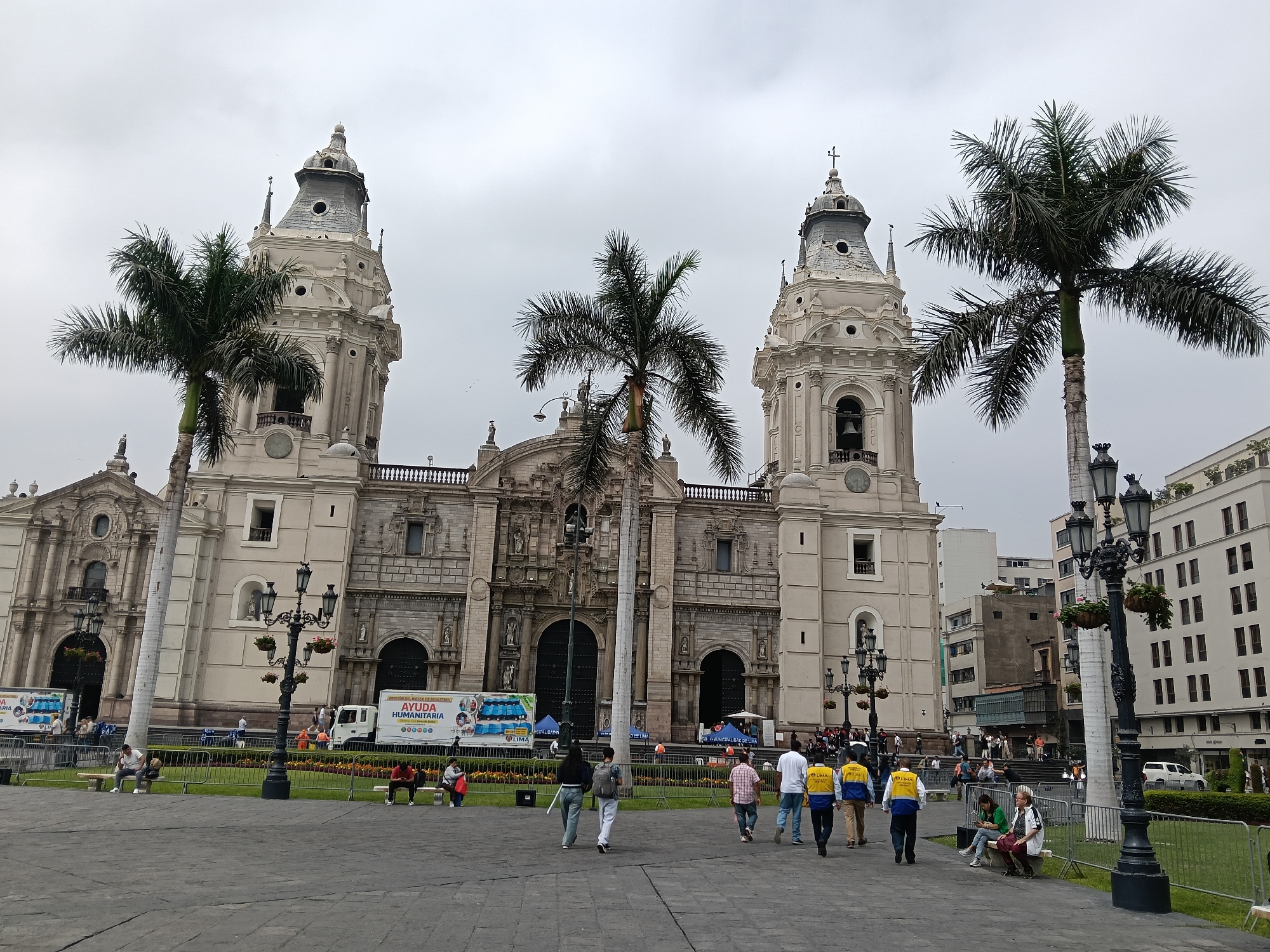 The image depicts the Plaza Mayor in Lima, Peru, with the Cathedral of Lima as the focal point. The cathedral is a large, ornate building with two tall towers. Palm trees stand in front of the cathedral and around the plaza. The sky is overcast. Several people are walking across the plaza. There is a truck parked near the cathedral displaying "AYUDA HUMANITARIA" (humanitarian aid) on its side. There are benches along the edge of the plaza where people are sitting. Street lamps are also visible.